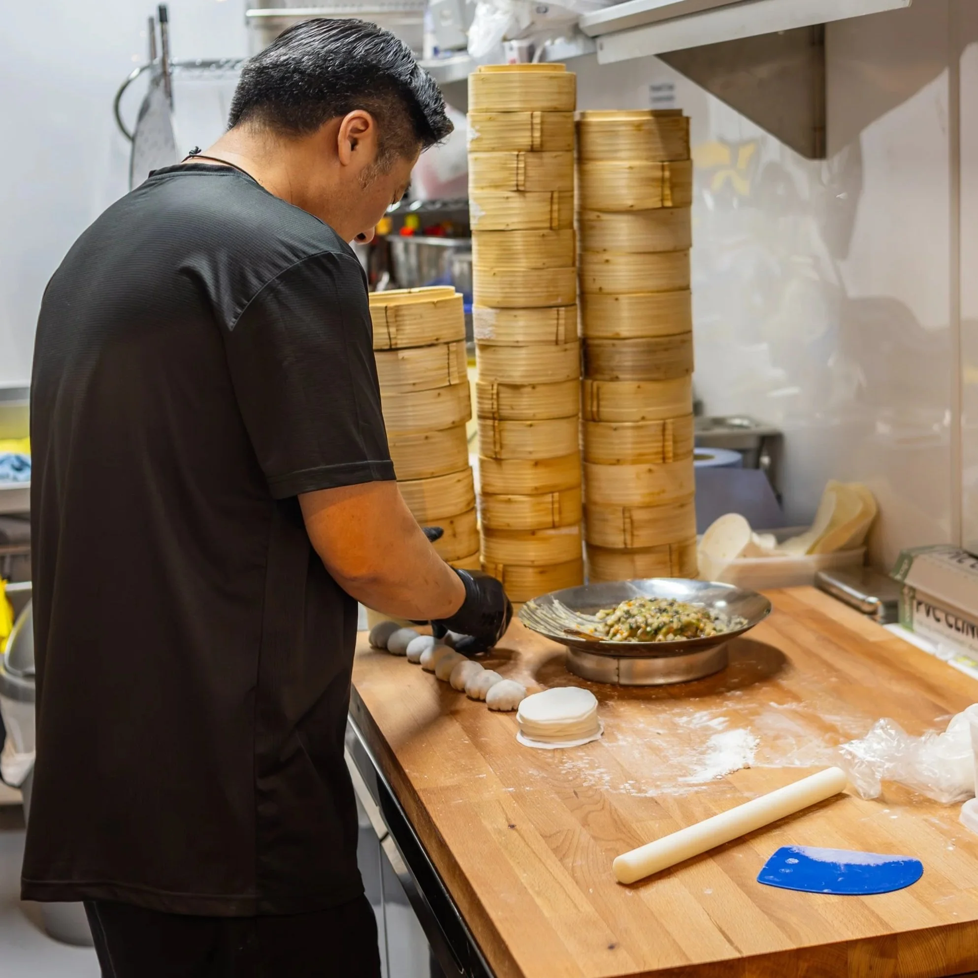 A chef preparing dumplings in a kitchen, with stacks of bamboo steamers on the counter and a bowl of filling nearby.