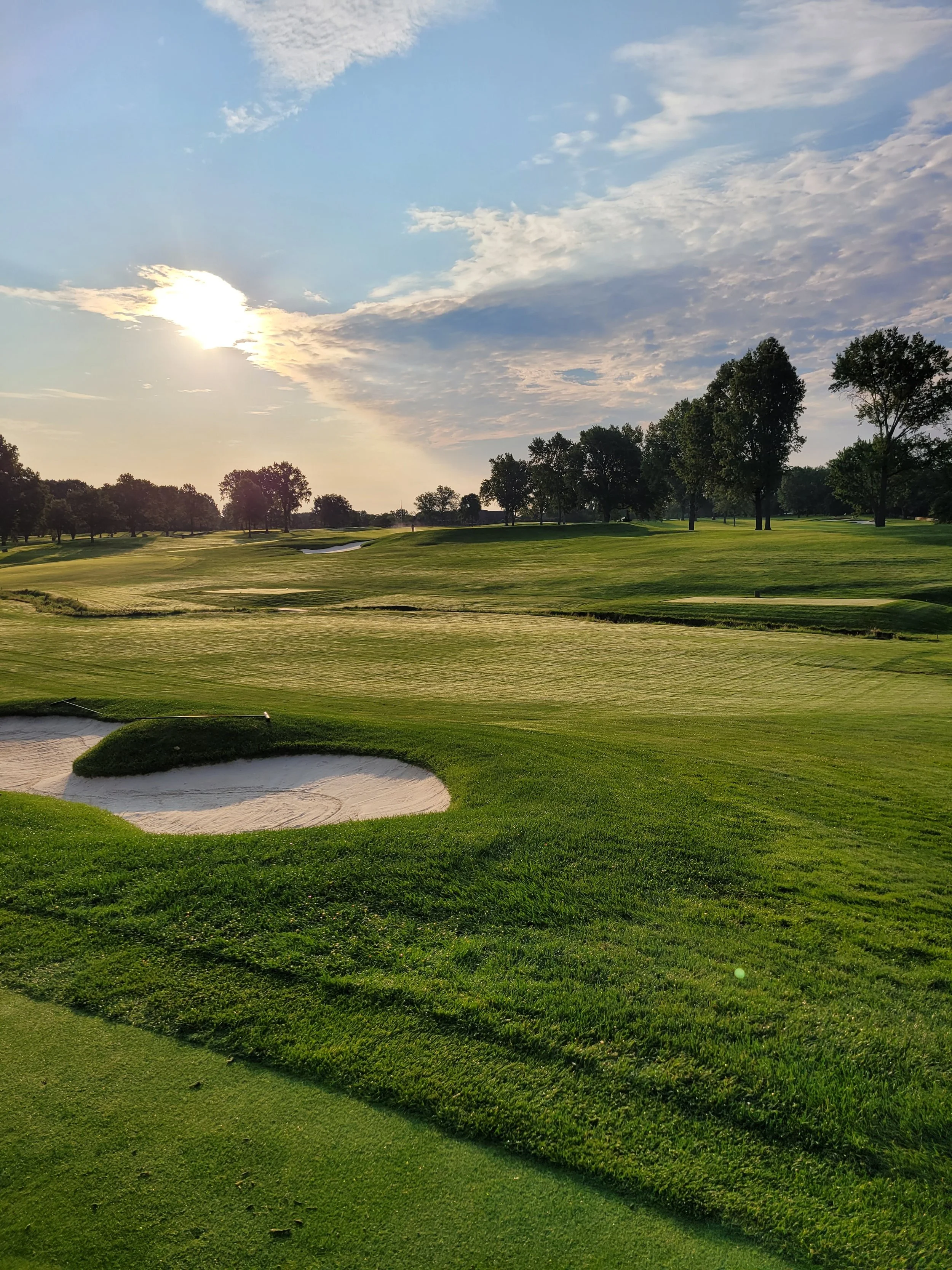 A scenic view of a golf course in the late afternoon with a green fairway, sand bunkers, tall trees, and a partly cloudy sky with the sun setting.