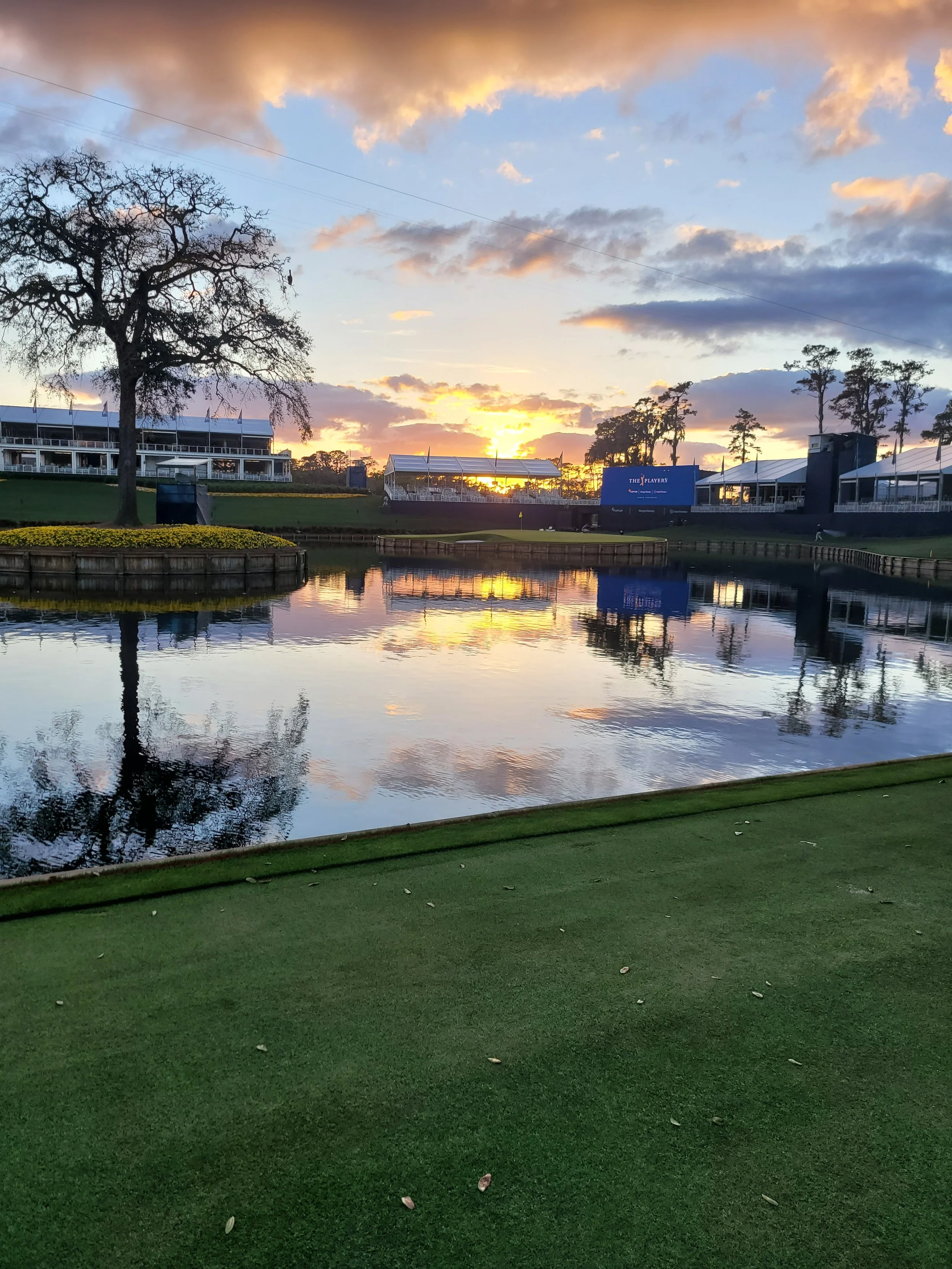 A golf course at sunset, with a small pond reflecting the colorful sky and trees around it.