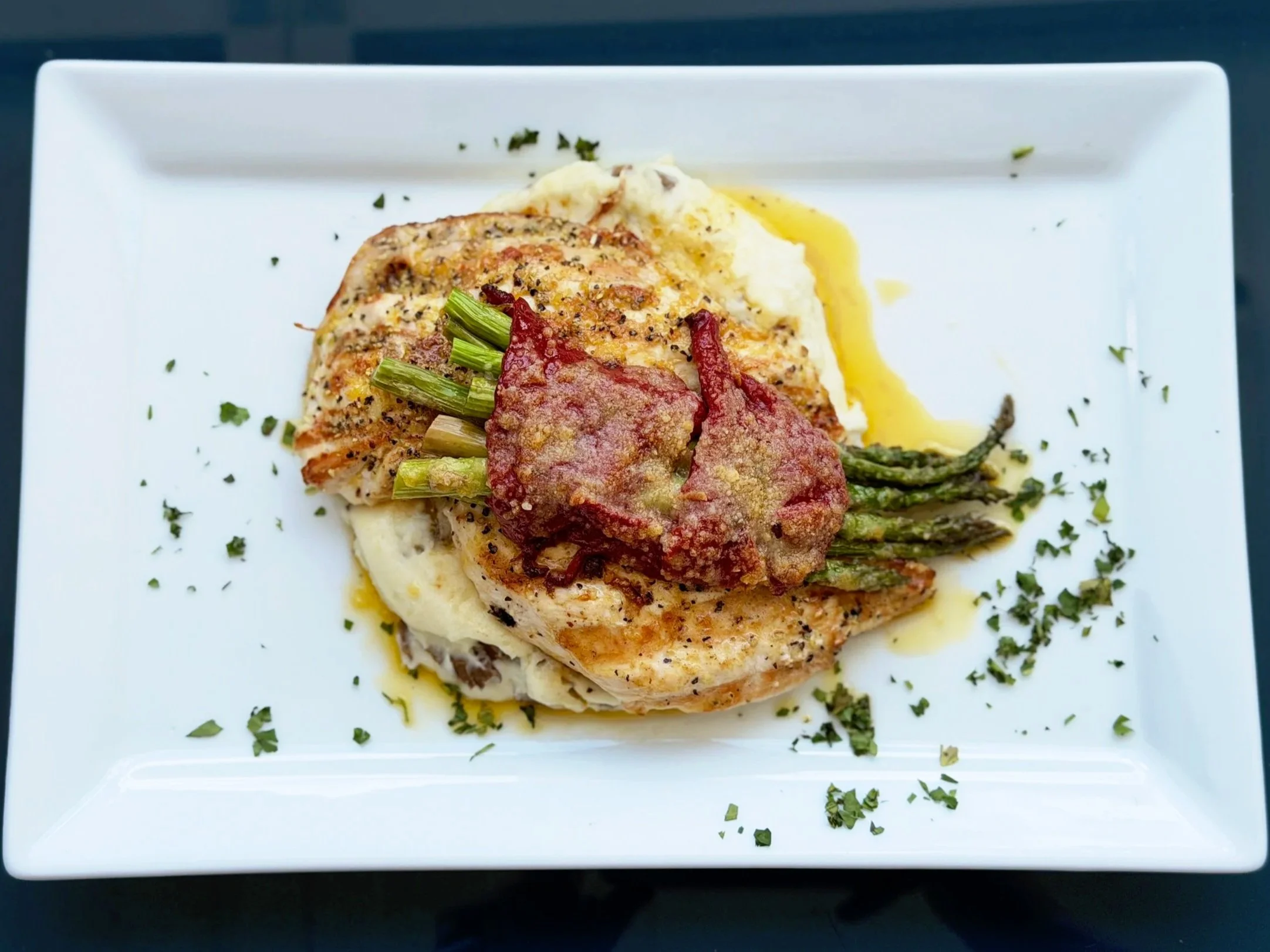 A plated dish featuring lemon pepper chicken topped with green herbs, served with mashed potatoes, grilled asparagus, and a slice of bacon, garnished with chopped herbs on a white square plate.