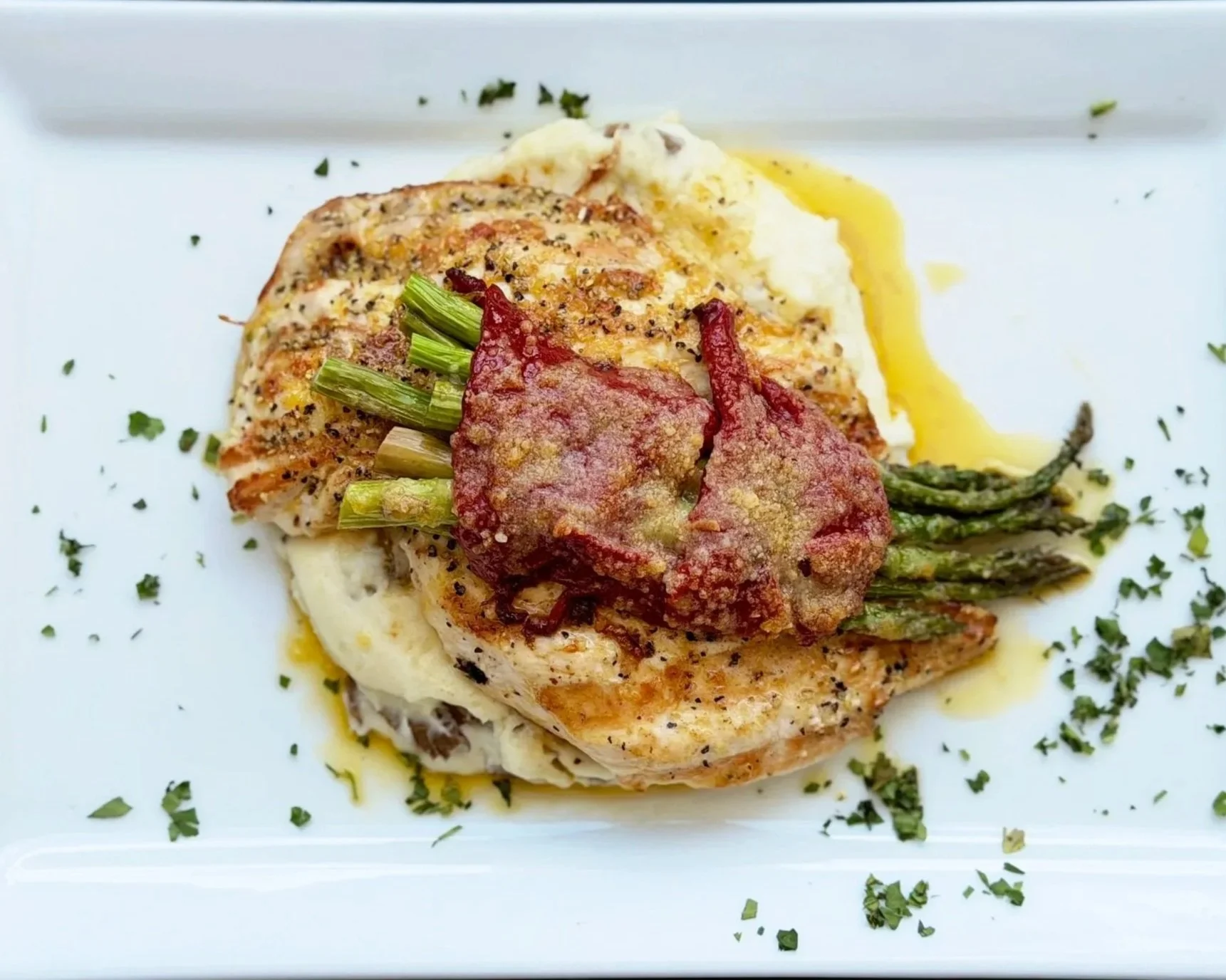 A plated serving of baked chicken topped with cream sauce, with asparagus spears and mashed potatoes, garnished with herbs.