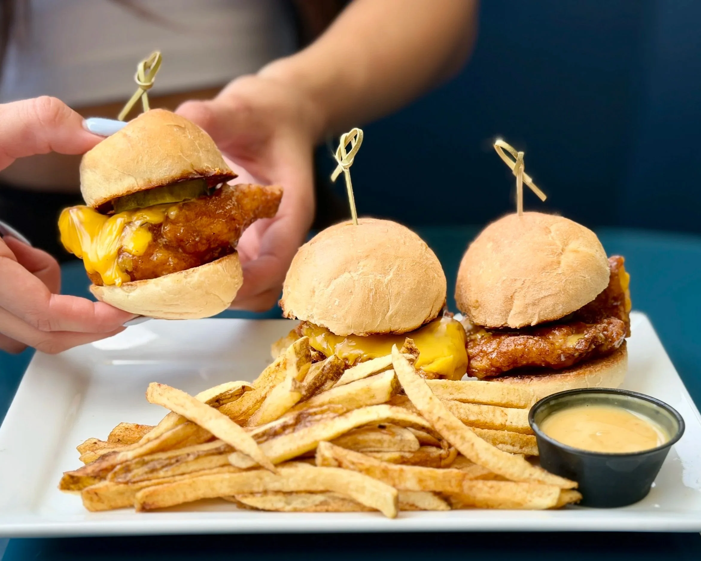 Close-up of a hand holding a mini sandwich with fried chicken, pickles, and mustard, with a row of similar sandwiches with skewers on a white tray and a small bowl of mustard in the background.