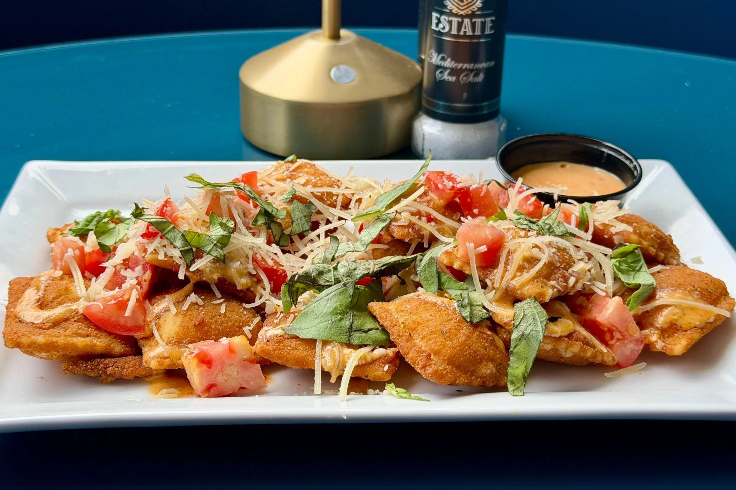 Fried fish fillet topped with chopped tomatoes, shaved Parmesan cheese, and fresh herbs, served with a side of dipping sauce, on a white rectangular plate.