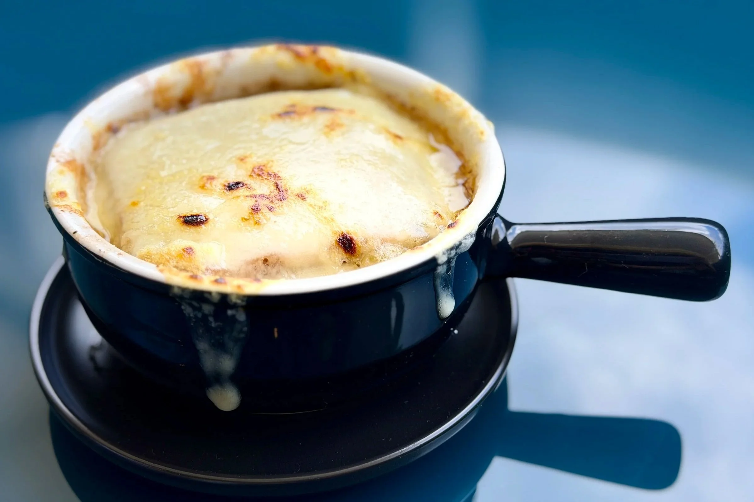 A black cup of baked French onion soup topped with melted cheese, placed on a matching black saucer.