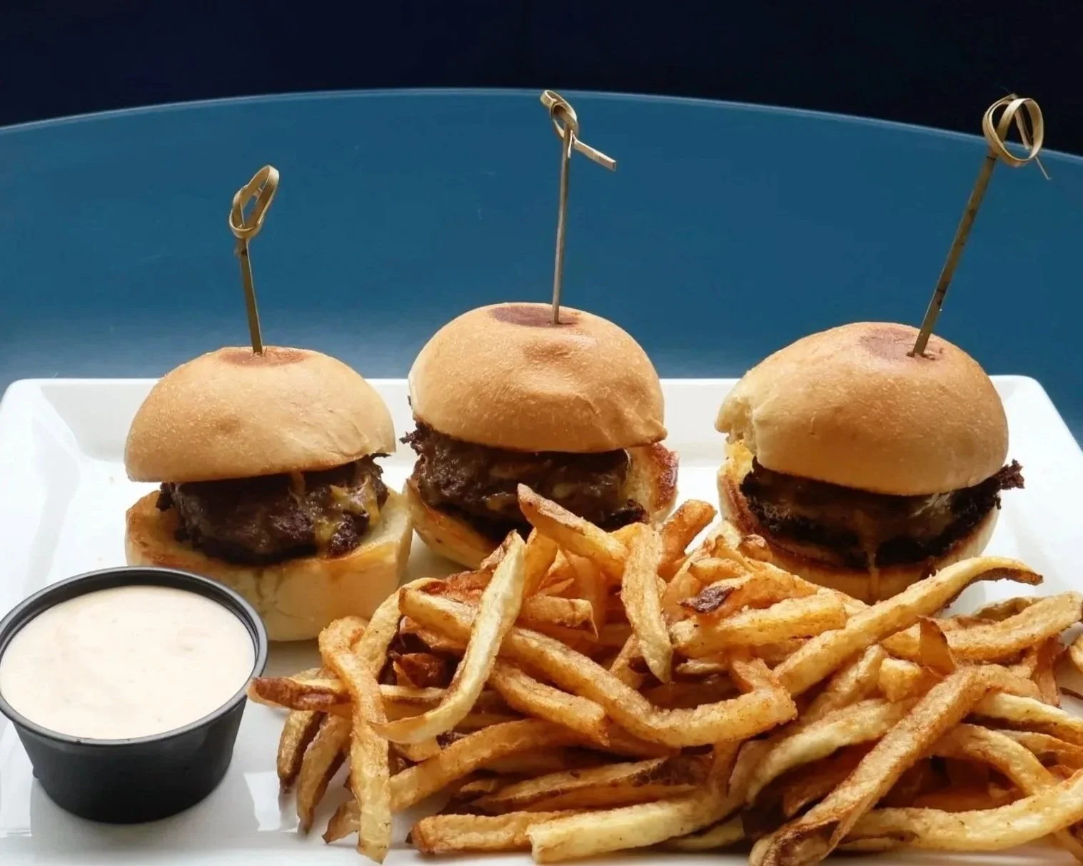 Three mini cheeseburgers with beef patties and buns on a white tray, served with French fries and a side of creamy dipping sauce.