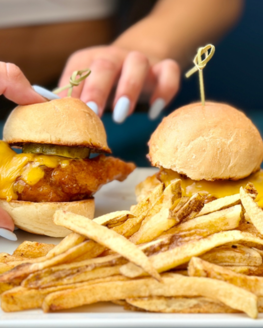 Close-up of two cheeseburgers with pickles and melted cheese, served with French fries on a white plate, with a person reaching for one burger.