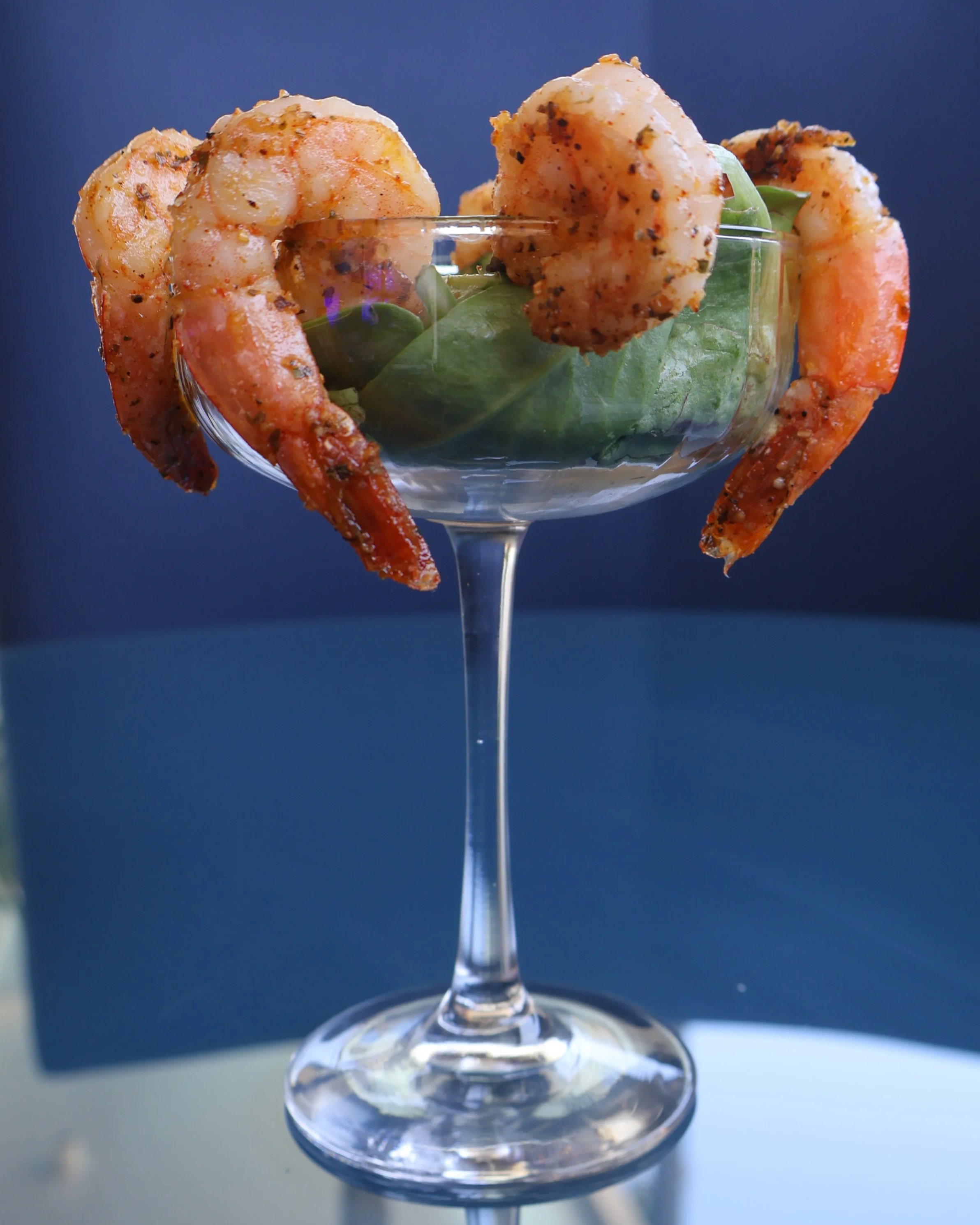 Close-up of a clear glass dessert dish with cooked shrimp garnished with seasoning, garnished with green leaves, against a dark blue background.