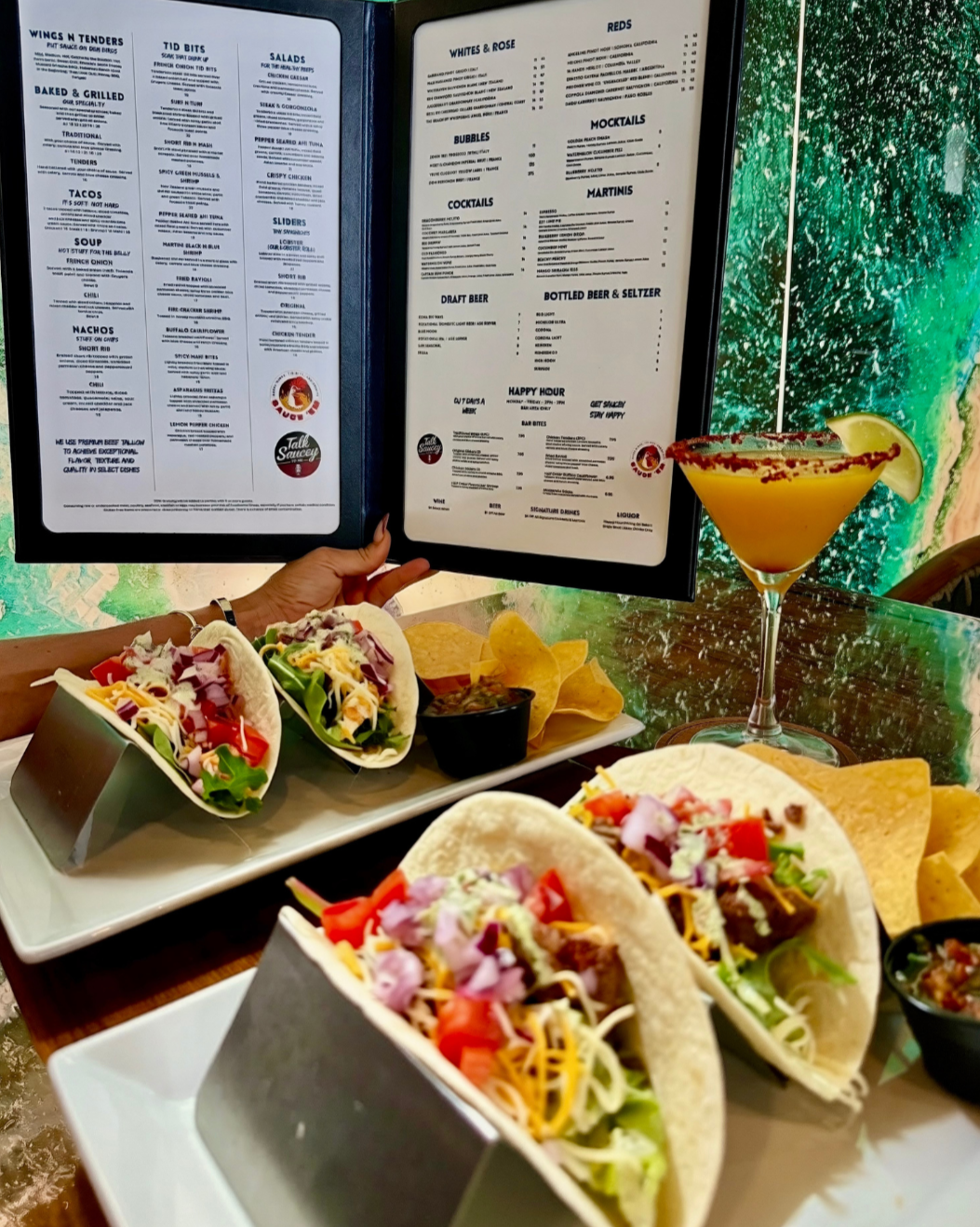 A restaurant table with three tacos filled with vegetables and shredded cheese, a side of chips with salsa, a cocktail with a lemon wedge, and a menu held by a person with a background of a green scenic wall.