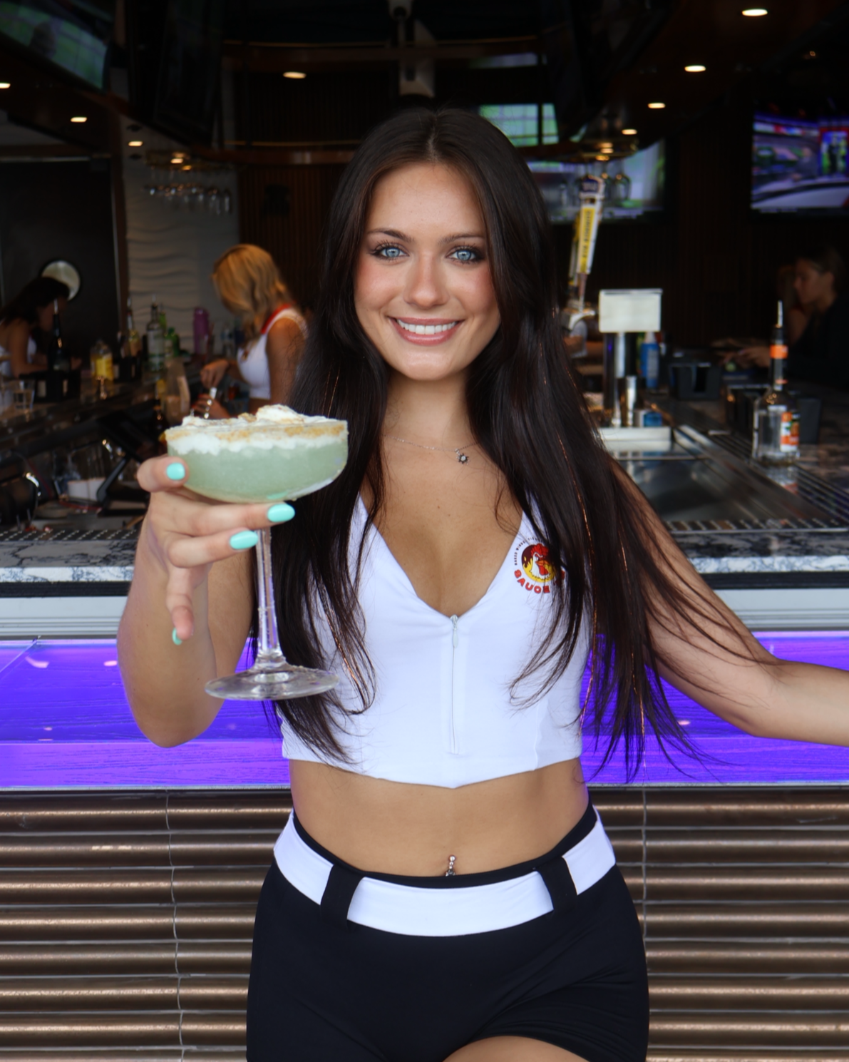 A young woman with long dark hair and blue eyes smiling and holding a cocktail in a bar, day setting. It is a key lime pie martini