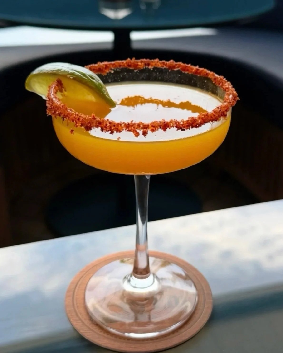 A cocktail in a coupe glass with a salted rim, garnished with a lime wedge, sitting on a wooden coaster.