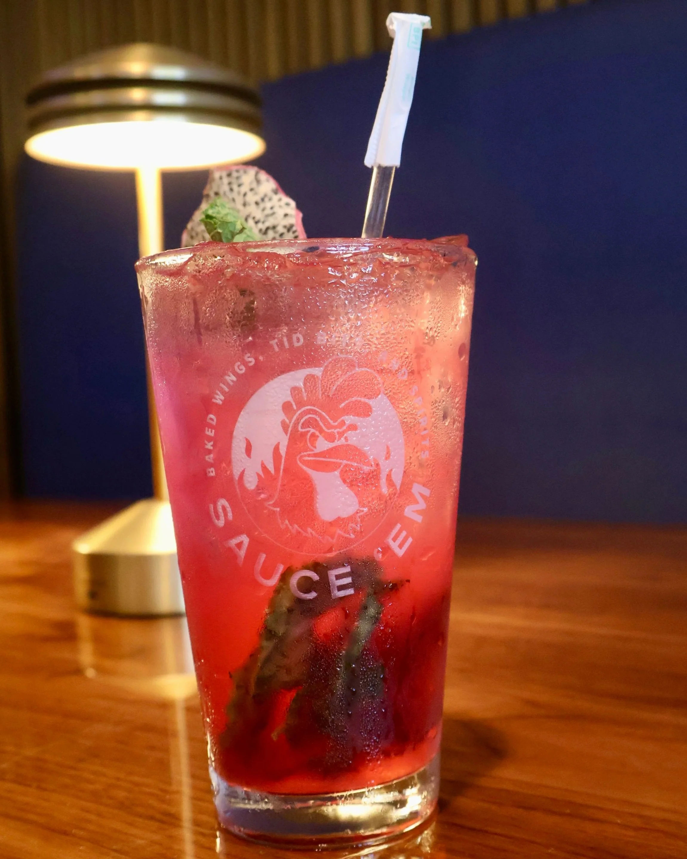 A tall glass of pink cocktail with ice, garnished with a slice of dragon fruit and a green leaf, next to a modern brass table lamp, on a wooden table with a dark background.