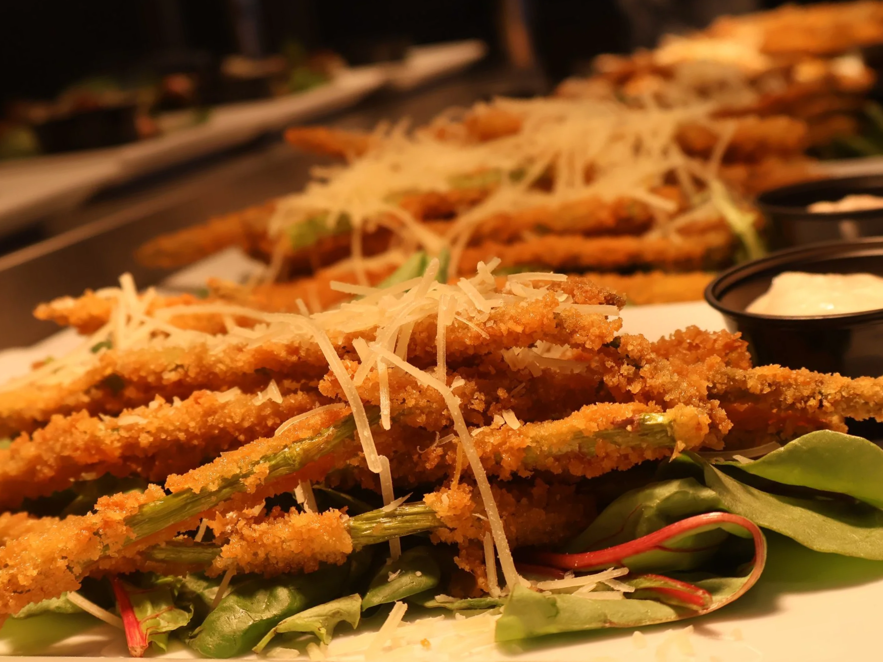 Close-up of fried asparagus spears topped with shredded cheese and served on a bed of mixed greens with small black containers of dipping sauce in the background.