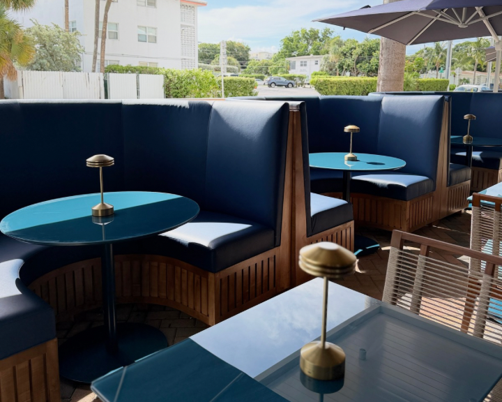 Outdoor seating area with blue booth seats, round blue tables, small metal lamps, and a large umbrella, with a view of trees and buildings in the background on a sunny day.