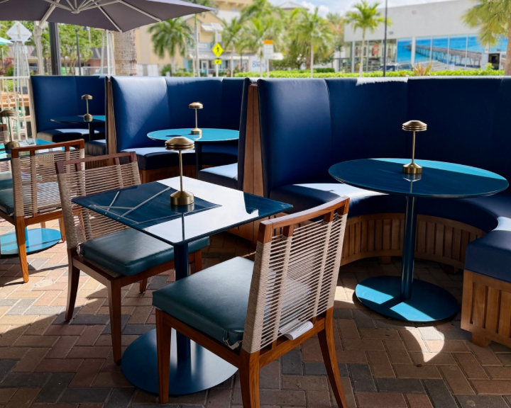Empty outdoor restaurant patio with blue booths, tables, and chairs, set up with small lamps, with palm trees and buildings in the background.