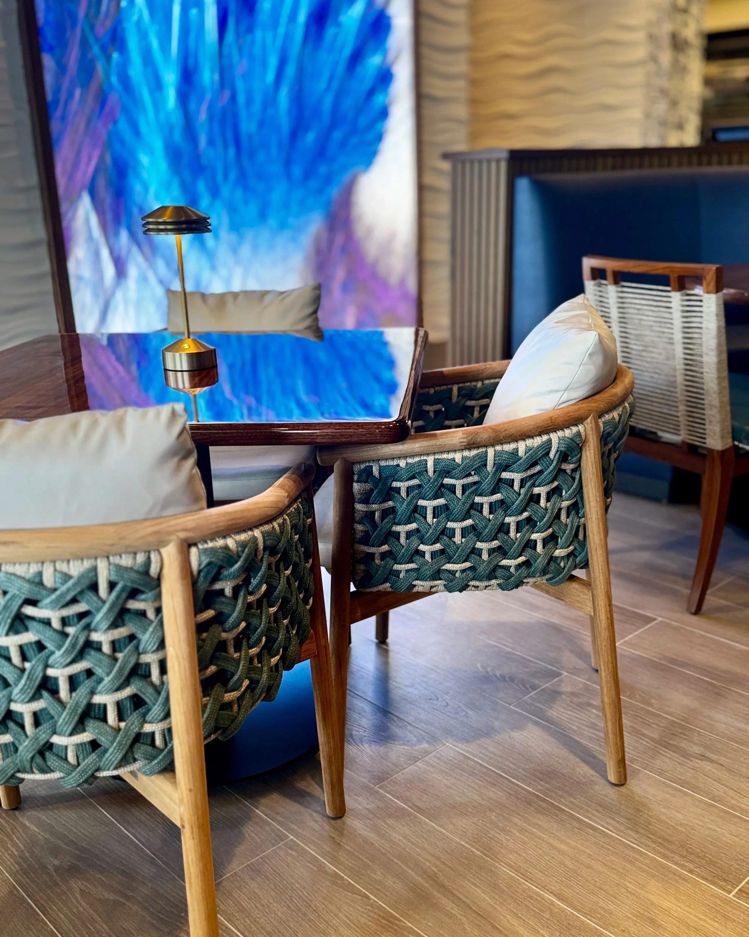A dining area with a wooden table and woven chairs. There is a modern table lamp with a gold base and black shades. The background features a blue abstract art piece and wood-paneled walls, with a section of blue upholstered paneling.