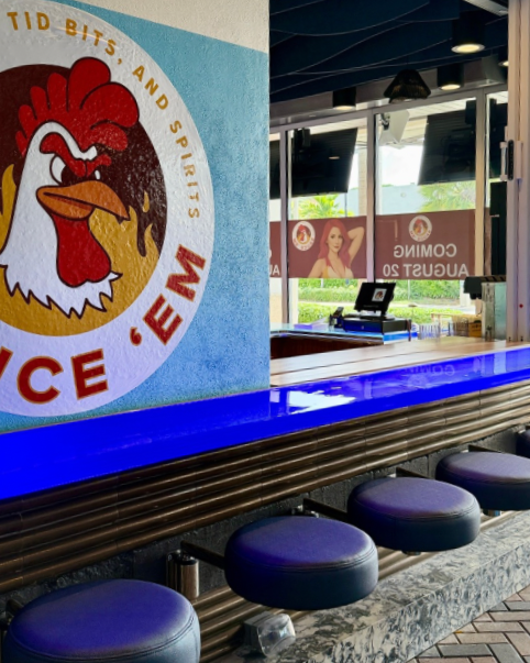 Interior of a restaurant or bar with a blue illuminated counter and black bar stools. There is a large graphic of a rooster with a red crest and yellow beak on the wall. Windows show outdoor seating and advertising banners.