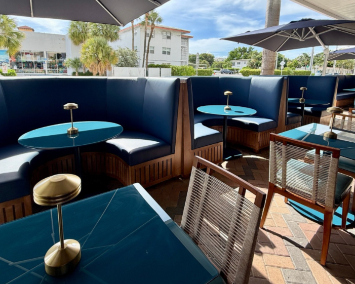 Outdoor seating area with blue booths, tables with small lamps, and umbrellas providing shade, in a sunny setting with palm trees and residential buildings in the background.