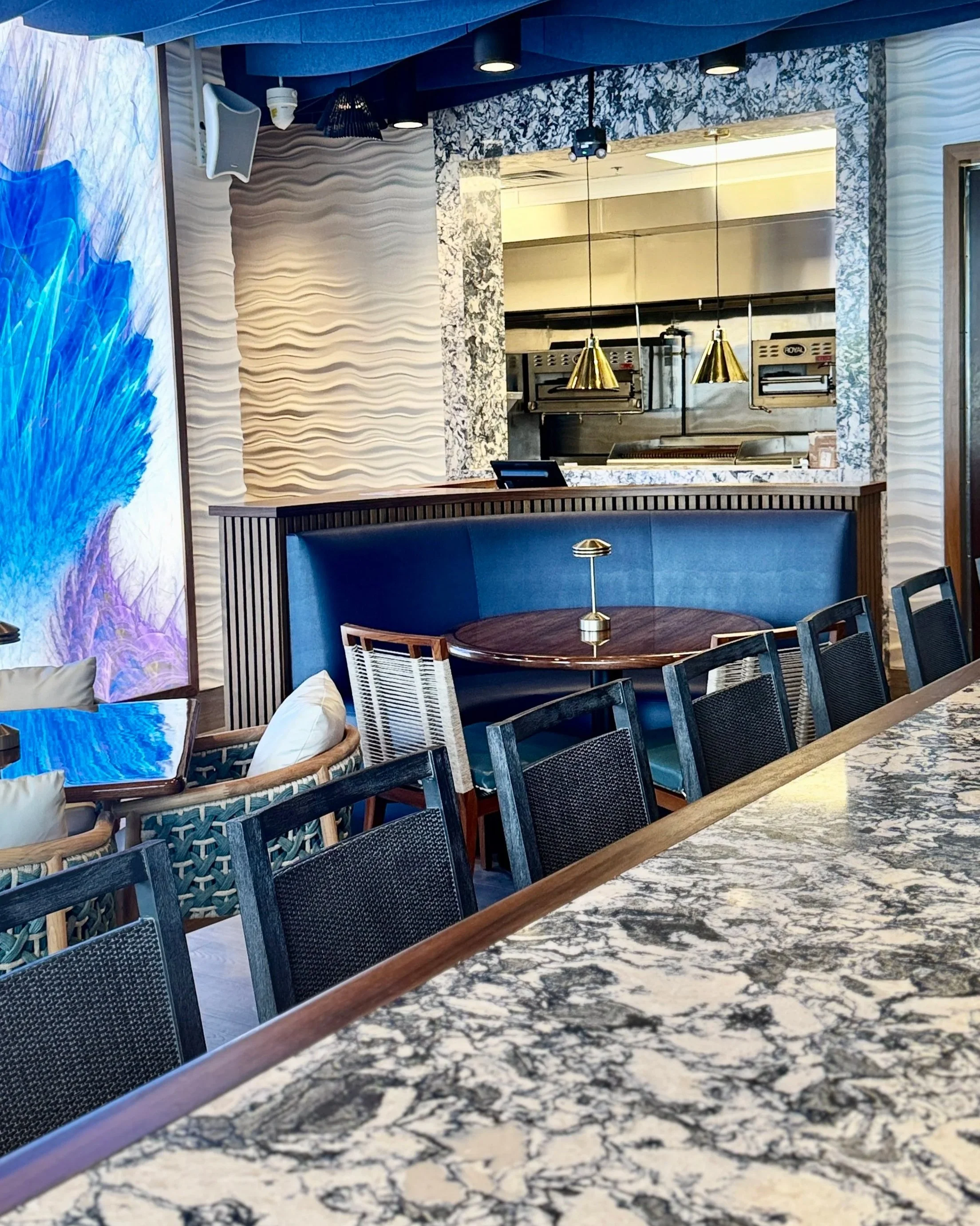 View of a modern restaurant interior with a marble countertop in the foreground, black chairs around a round wooden table with a small lamp, patterned wall panels, gold pendant lights, and a mirror reflecting a kitchen area with stainless steel appli