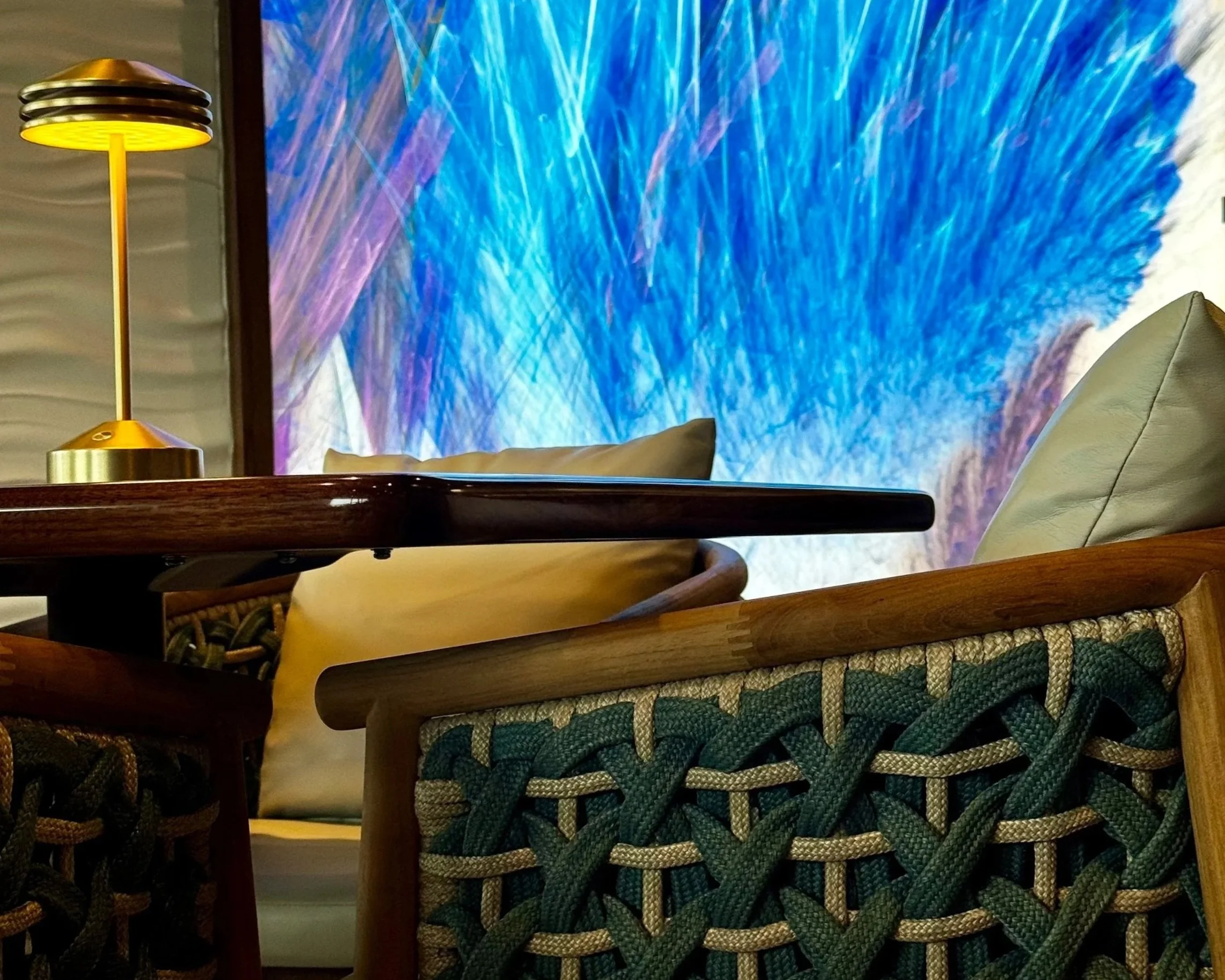 The image shows a wooden table in a living room, with a modern yellow and silver table lamp, a beige sofa, a colorful abstract wall art piece with shades of blue, purple, and white, and woven basket chairs with green and white patterns.