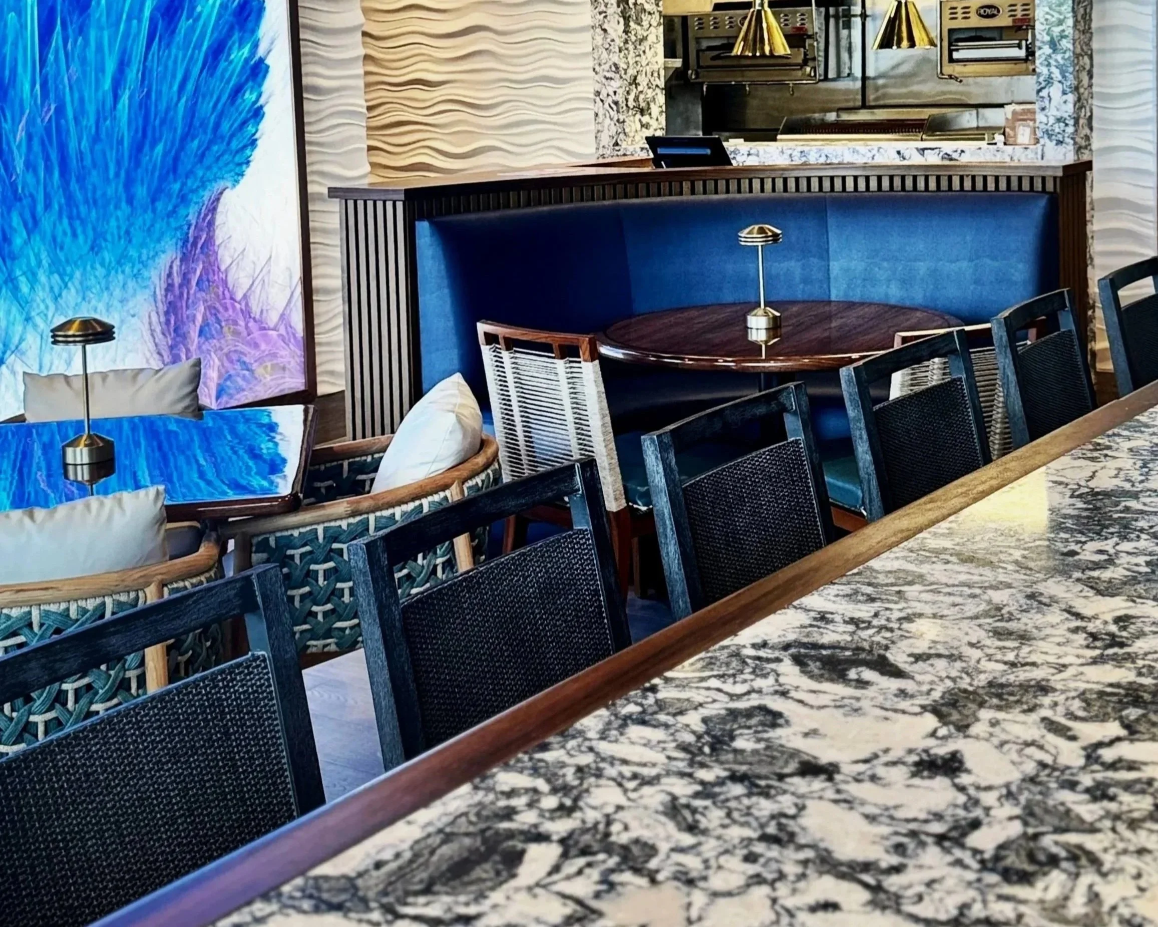 A restaurant or cafe interior with a round wooden table, black chairs, blue cushioned bench seating, a marble countertop, decorative golden pendant lights, and textured wall panels.