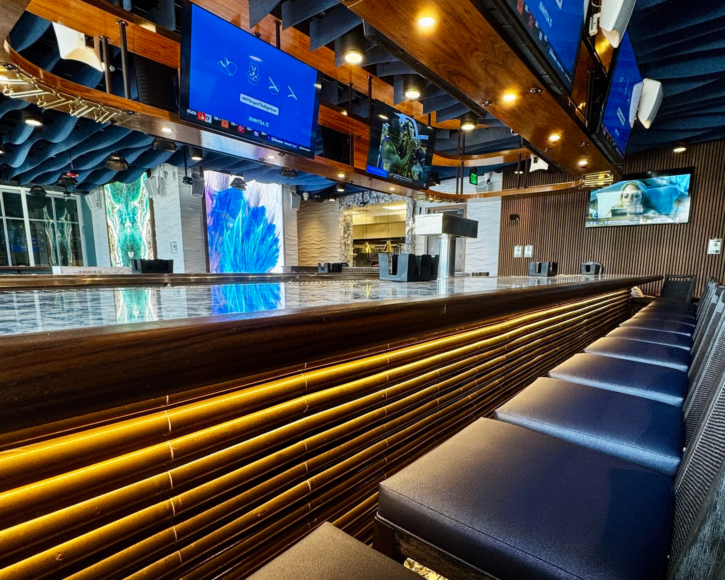 Interior of a modern bar or restaurant with multiple mounted flat-screen TVs, a wooden ceiling with built-in lighting, a marble-topped bar counter, and dark-colored seating.