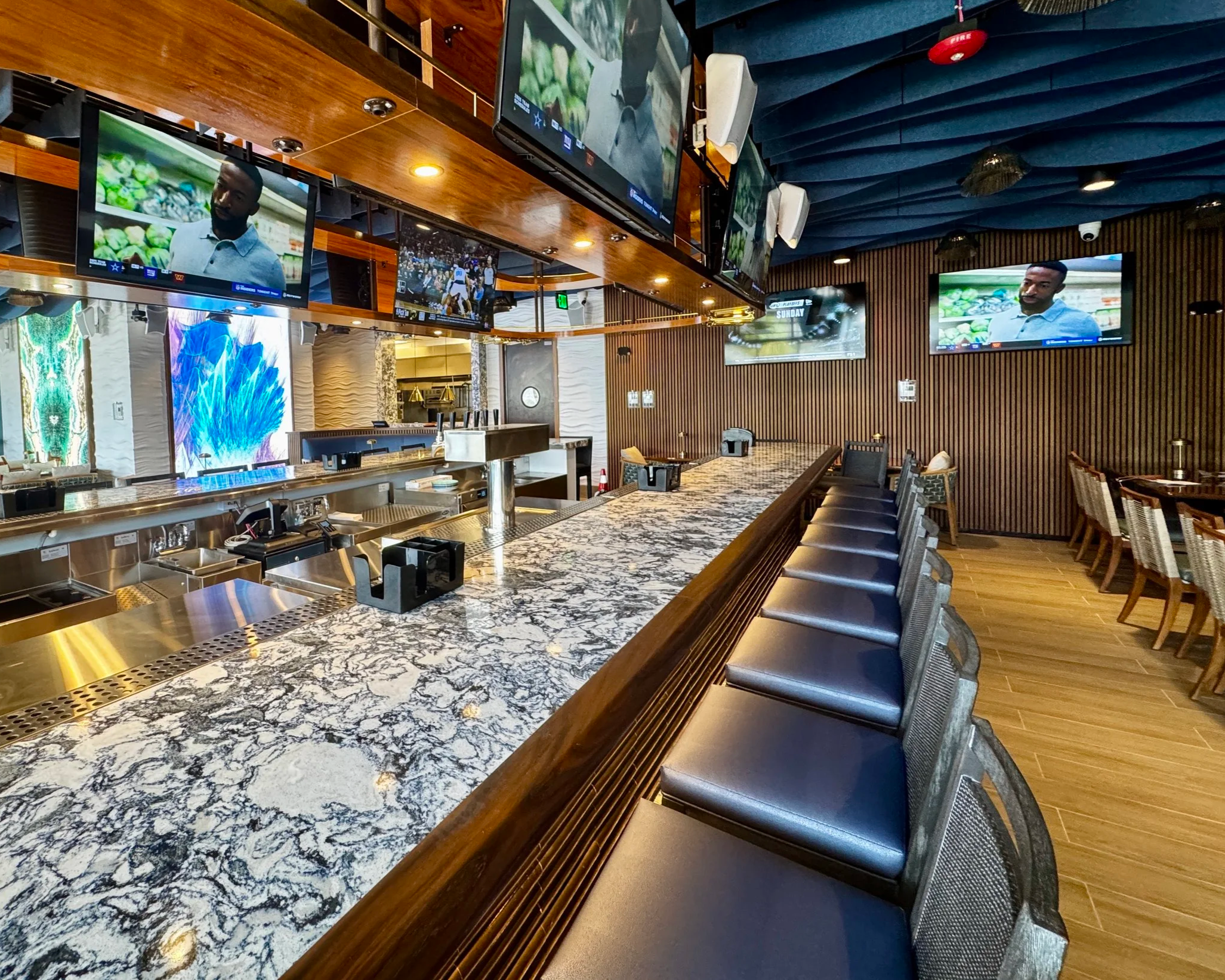 View of a modern bar area with a marble countertop, black bar stools, and multiple flat-screen TVs mounted on a wooden and blue-decorated wall.