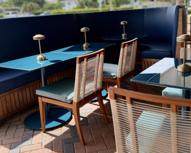 Empty outdoor seating area with tables and chairs, featuring blue and glass tabletops with small lamps, on a brick patio with a view of greenery.