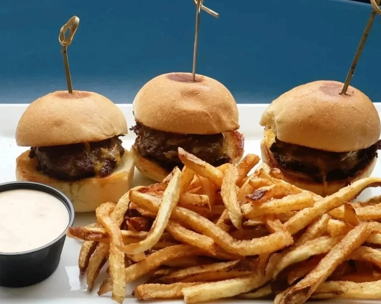 Three mini cheeseburgers with beef patties and buns on a white tray, served with French fries and a side of creamy dipping sauce.