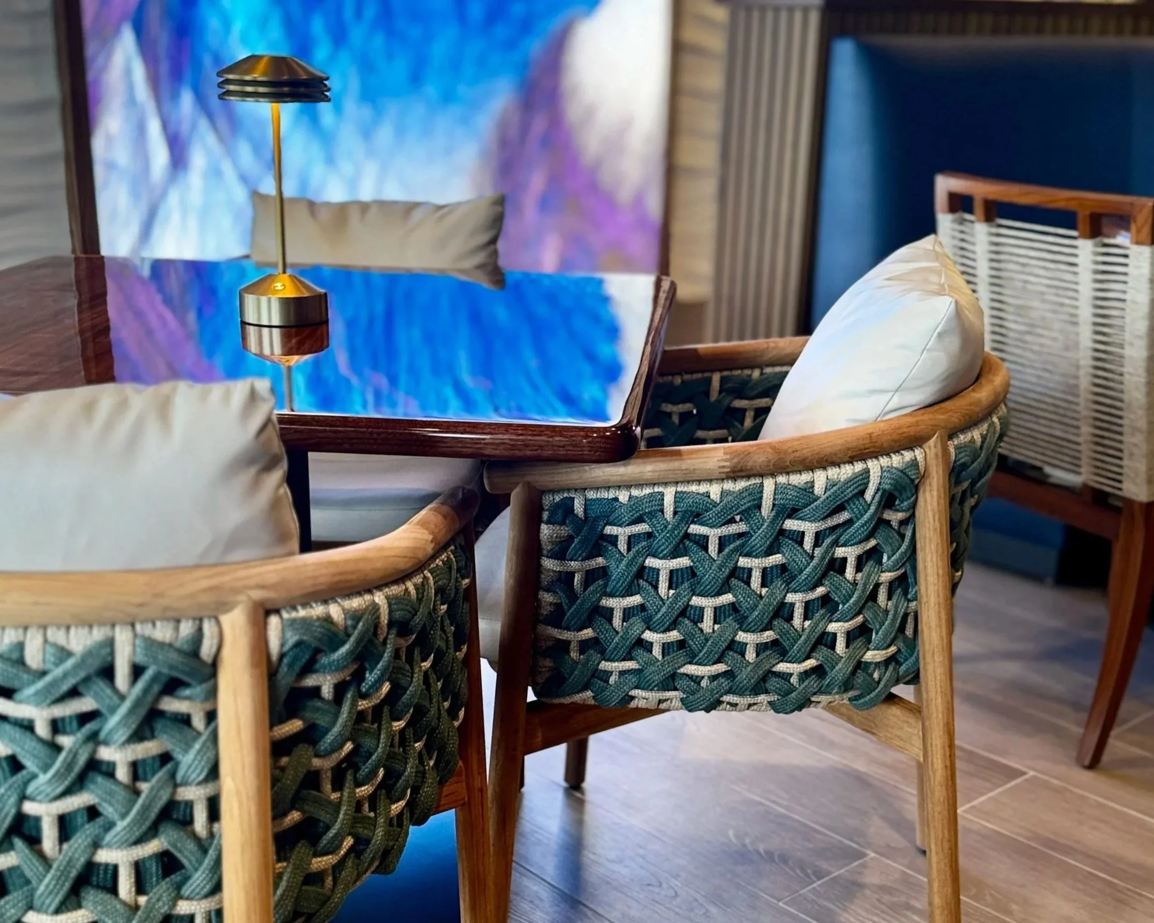 A dining area with a wooden table and woven chairs. There is a modern table lamp with a gold base and black shades. The background features a blue abstract art piece and wood-paneled walls, with a section of blue upholstered paneling.