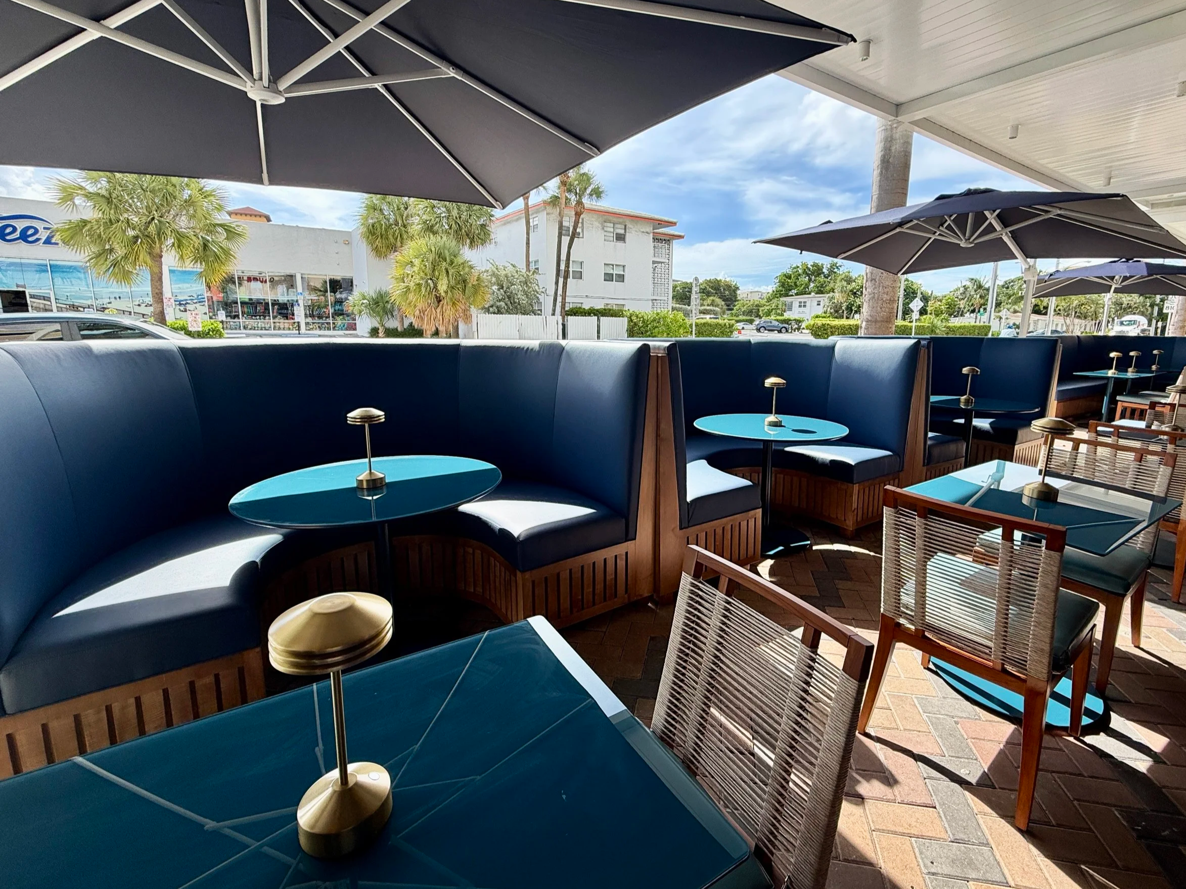 Beautiful patio outdoor seating area with blue booths, small round tables with gold candlestick holders, and wooden chairs under large gray umbrellas, overlooking a street with palm trees and white buildings.