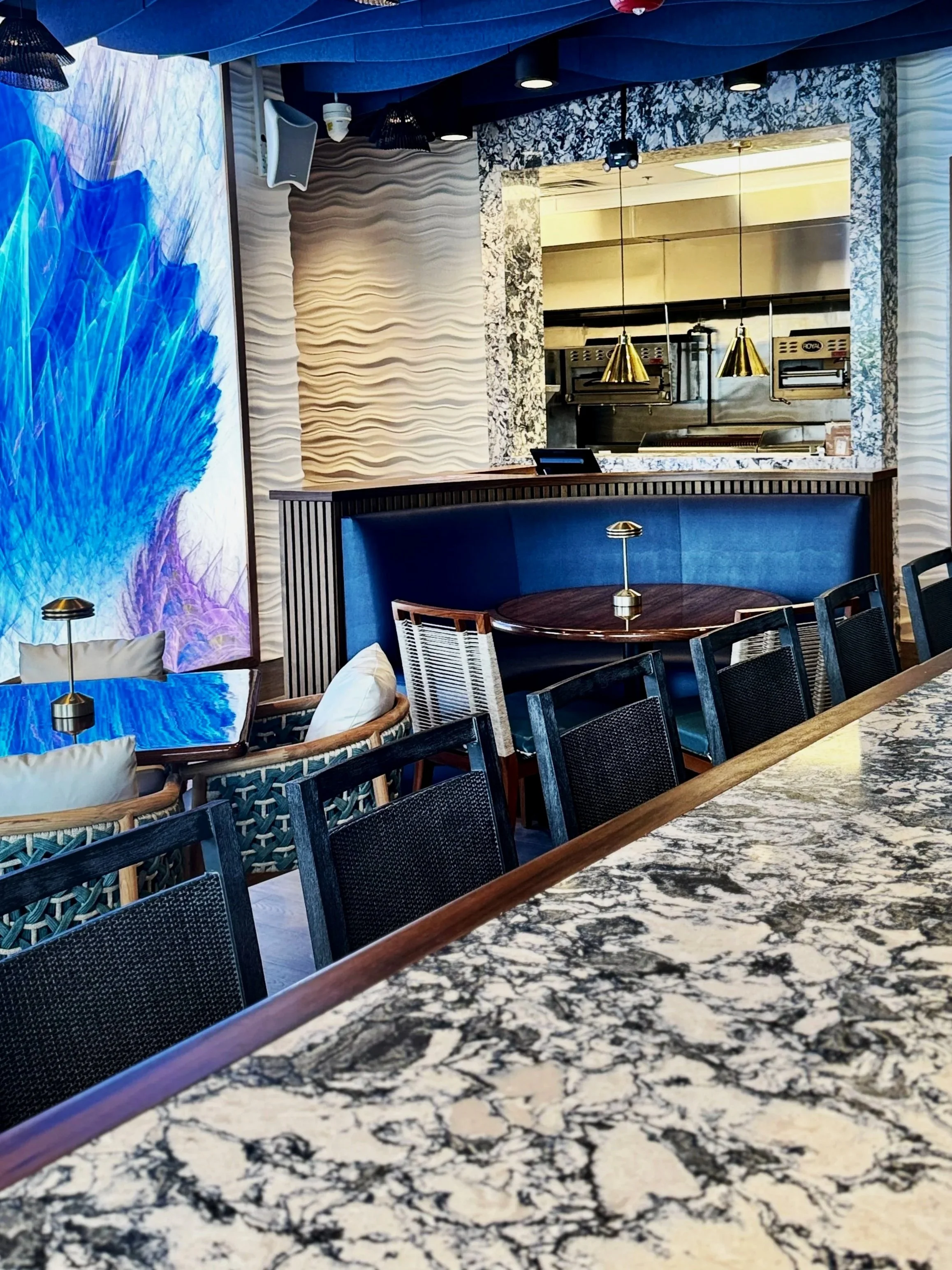 Interior of a modern restaurant with patterned walls, a marble table in the foreground, and a view into the open kitchen with hanging gold pendant lights.