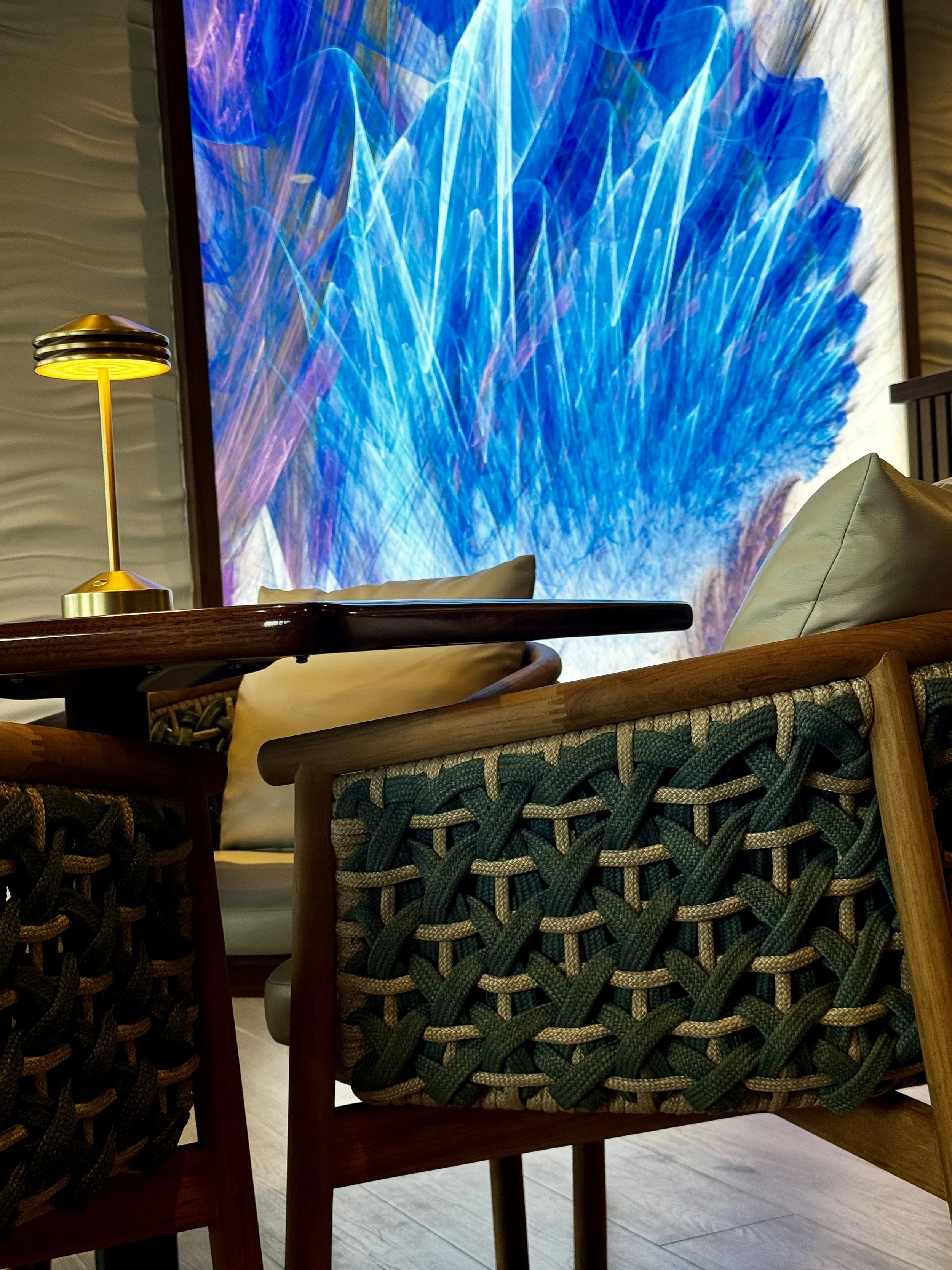 Close-up of a wooden chair with a woven green and beige seat, behind a glass table in a room with a large blue abstract artwork on the wall, a modern yellow desk lamp, and beige cushions.