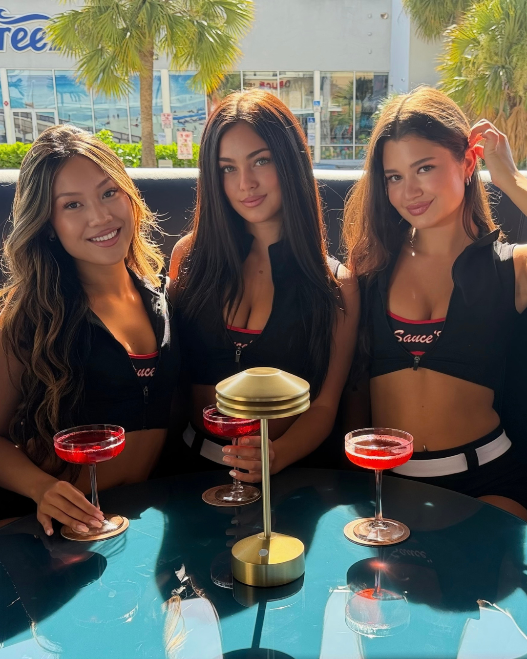 Three women sitting at a table with red drinks, smiling, with palm trees and a building in the background. Deerfield beach, FL in background