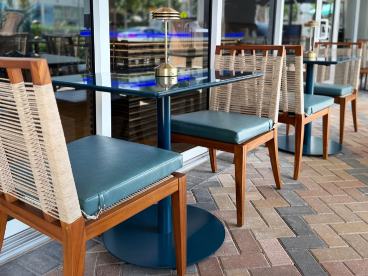 Empty outdoor dining table with blue base and a brass table lamp, surrounded by wooden chairs with woven backs and blue cushions, on a brick patio.