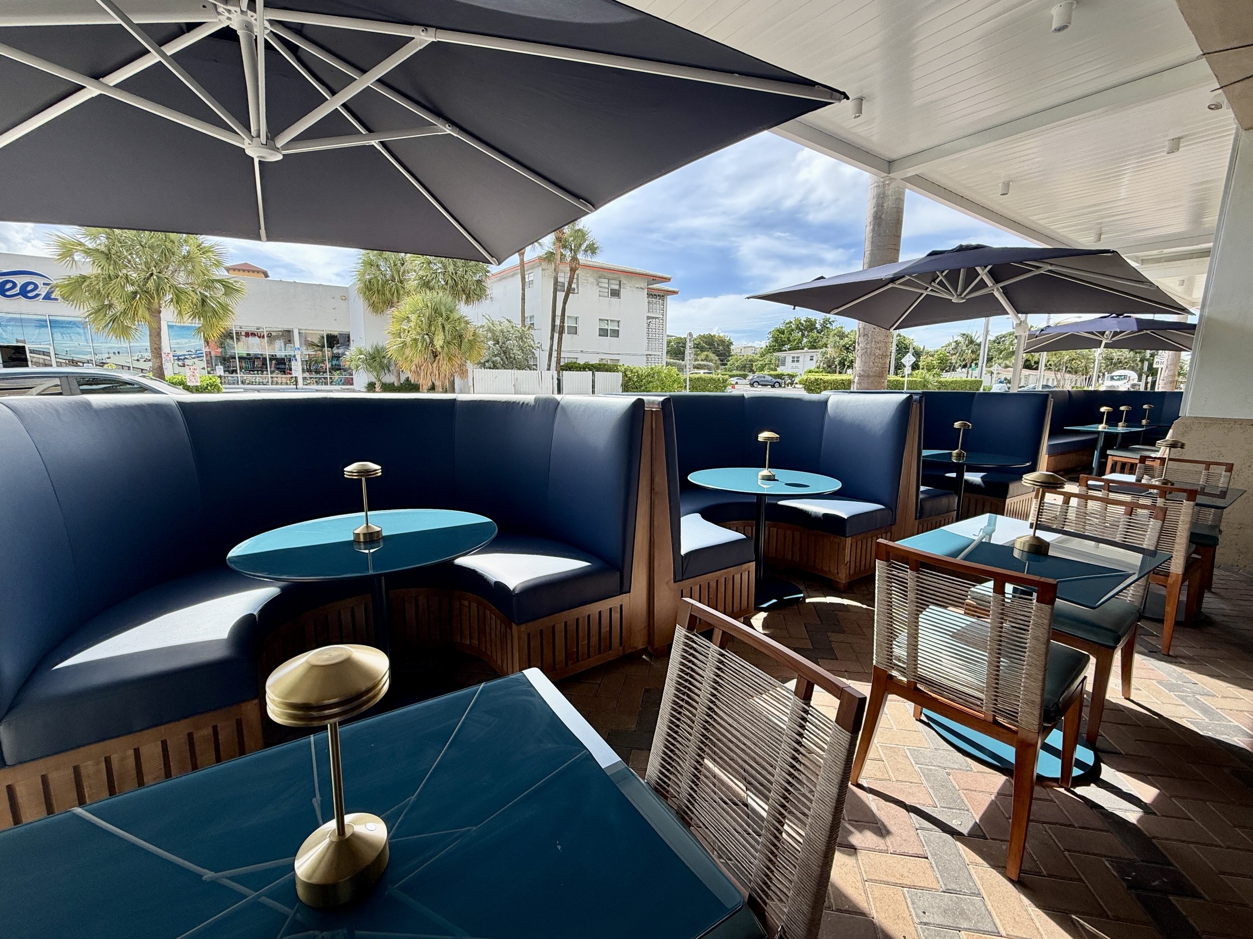 Outdoor patio area with blue cushioned booth seating, small blue tables with lamps on top, and wood chairs. Large umbrellas provide shade. In the background, palm trees and buildings are visible under a partly cloudy sky.