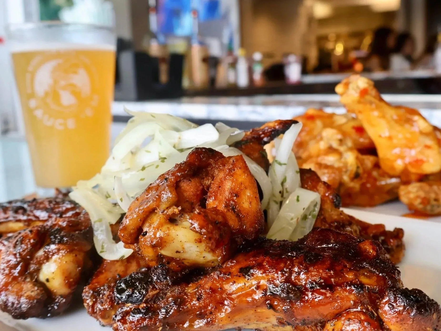 grilled chicken wings, sweet chili, hot tangy barbecue sauce, served with shredded lettuce and a slice of grilled sausage on a plate. In the background, a glass of beer with a logo, blurred restaurant interior with people and colorful decor.