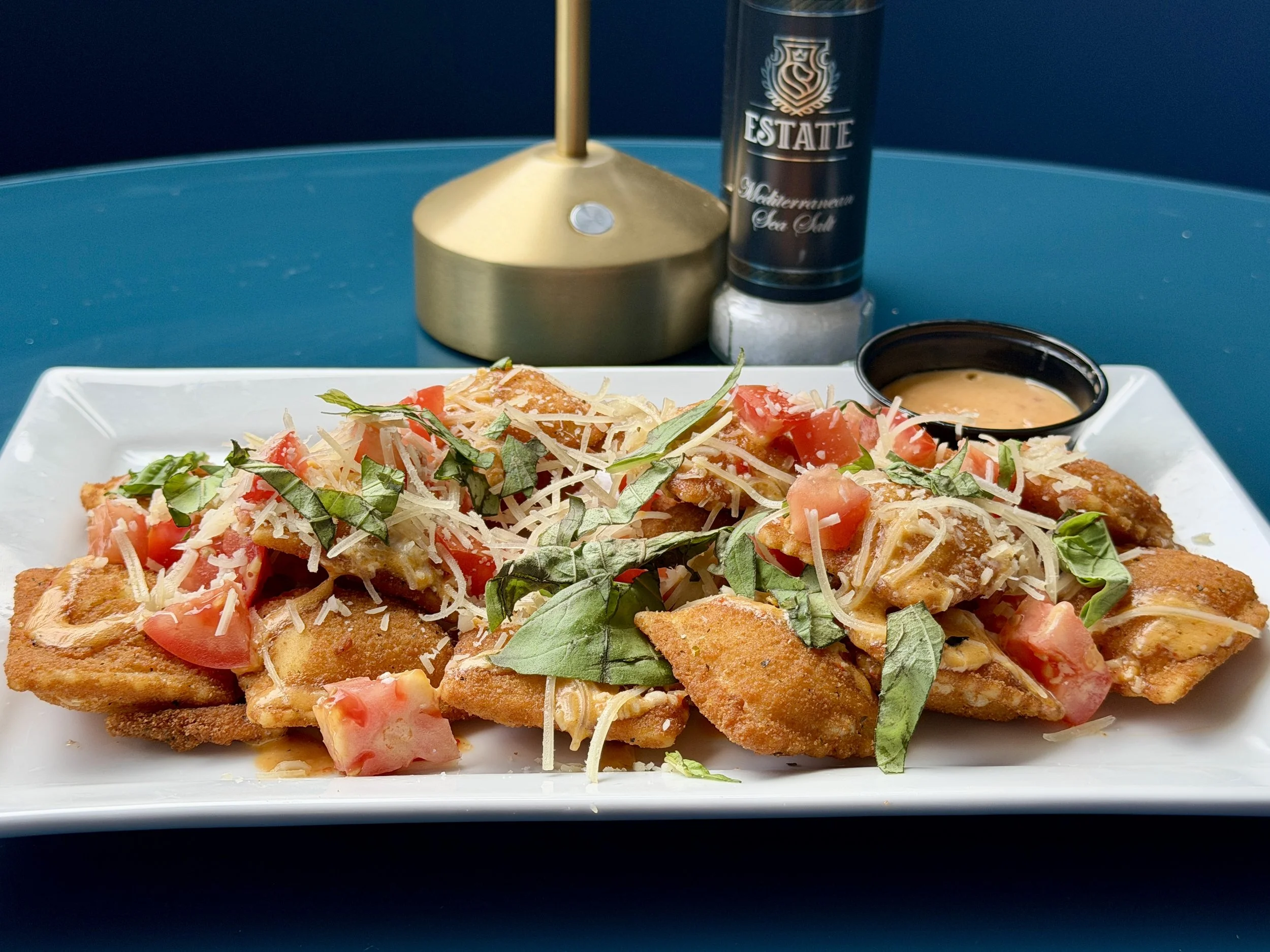 Close-up of a fried ravioli topped with chopped tomatoes, shredded cheese, and fresh basil, served on a white plate with a side of dipping sauce. spicy three-pepper blue cheese