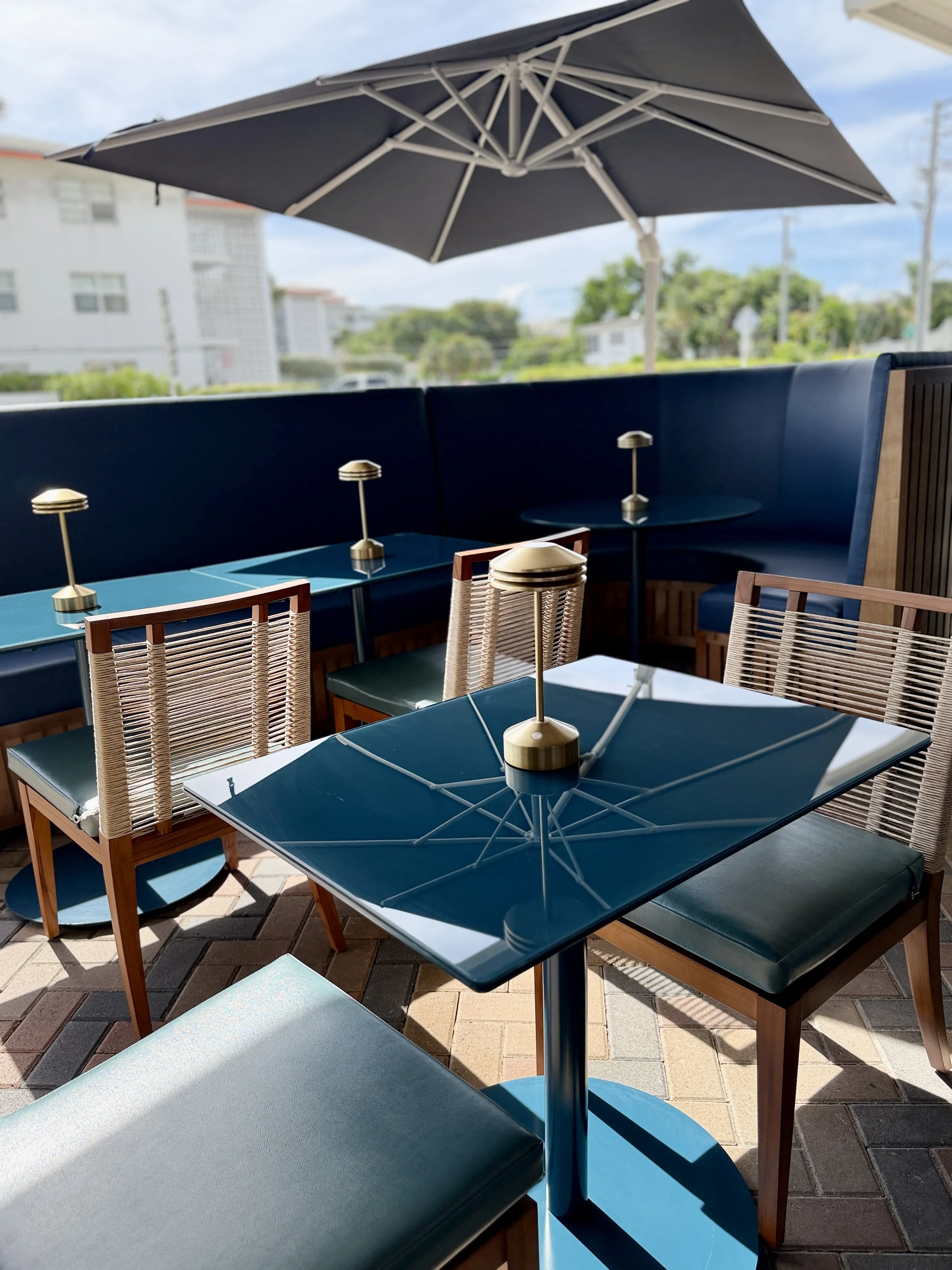 Outdoor patio seating area with blue tables, chairs, and a large umbrella, with buildings and trees in the background.