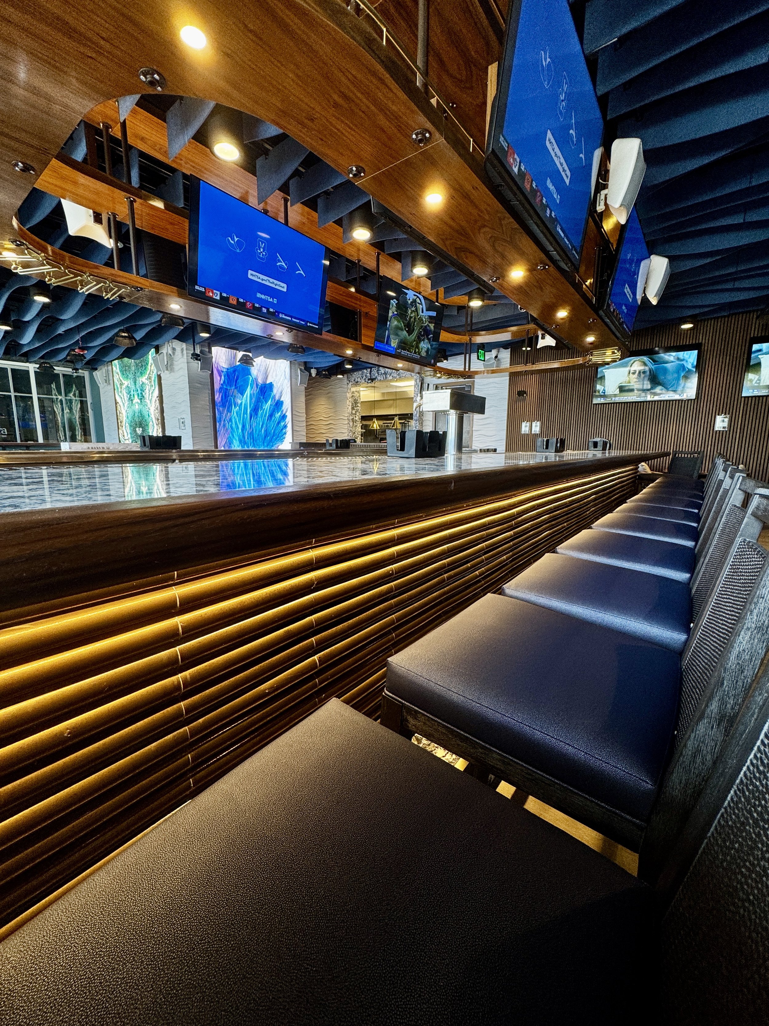 A modern bar interior with a curved wooden bar counter, black cushioned bar stools, multiple overhead television screens, and decorative lighting, with a textured wall and large window in the background.