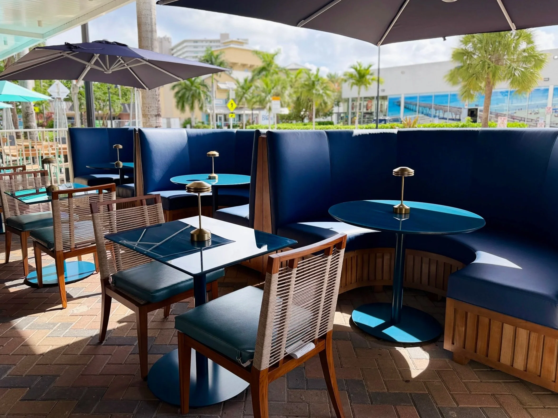 Outdoor seating area with blue upholstered booths, wooden chairs, and black tables under large umbrellas, surrounded by palm trees and storefronts.