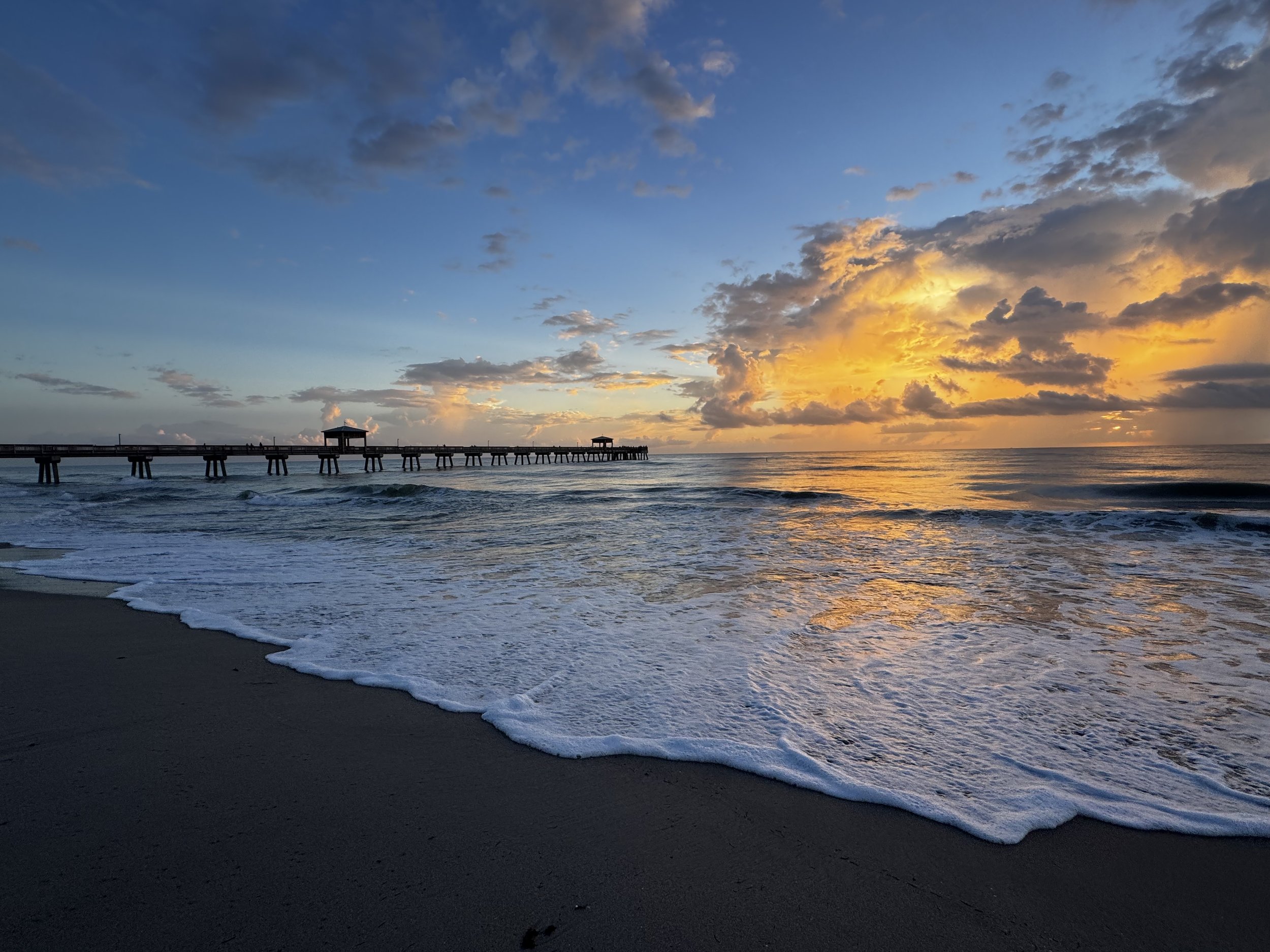 Sunset over the ocean with a pier extending into the water and clouds in the sky.