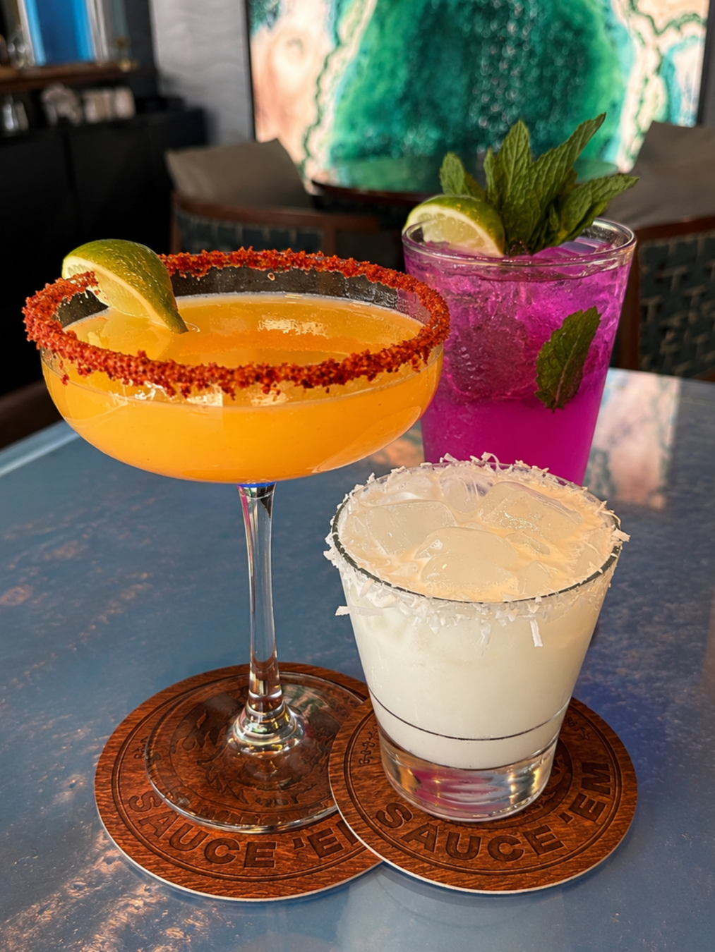 Three colorful cocktails on a table. The left cocktail is a mango habanero margarita with lime wedge and tajin rim, the middle one is pink blueberry lemonade with blueberry vodka with mint and lime, and the right one is white with a salted rim.