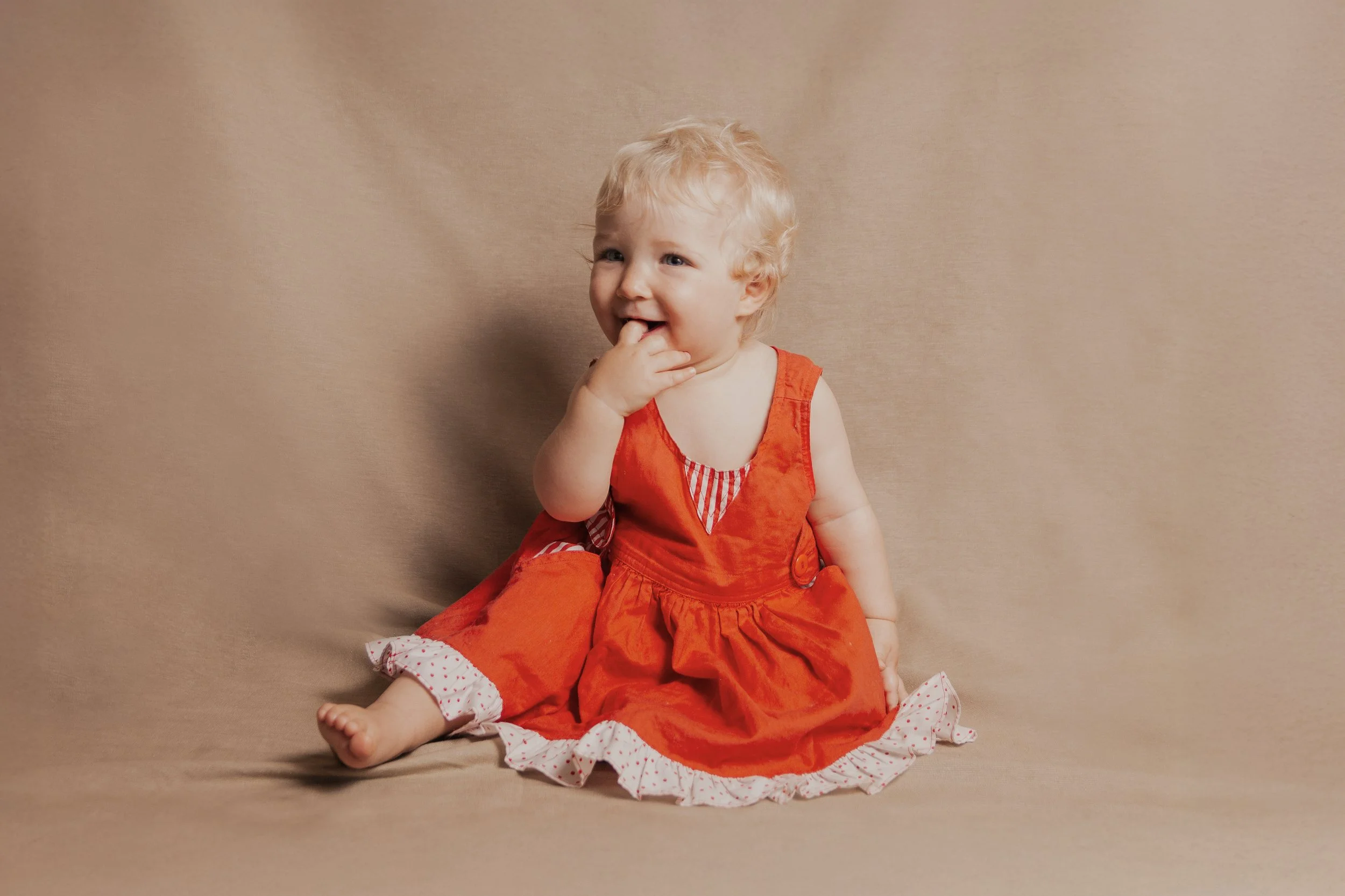Ein lachendes kleines Kind mit blondem Haar sitzt auf einem beigefarbenen Hintergrund, trägt ein rotes Kleid und schaut neugierig.