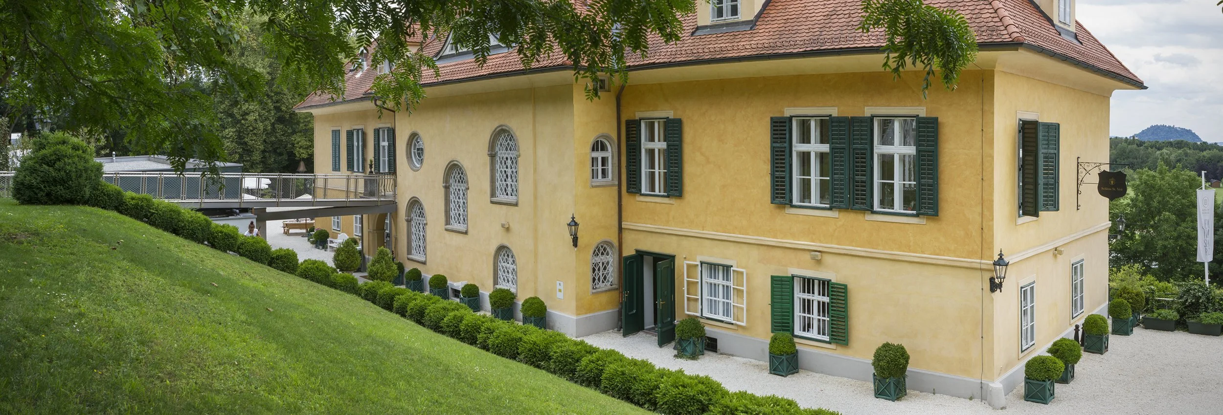 Buntes Haus mit gelber Fassade, grünen Fensterläden und roten Ziegeldach, umgeben von gepflegtem Garten mit Büschen und Bäumen, in ländlicher Umgebung unter blauem Himmel mit Wolken.