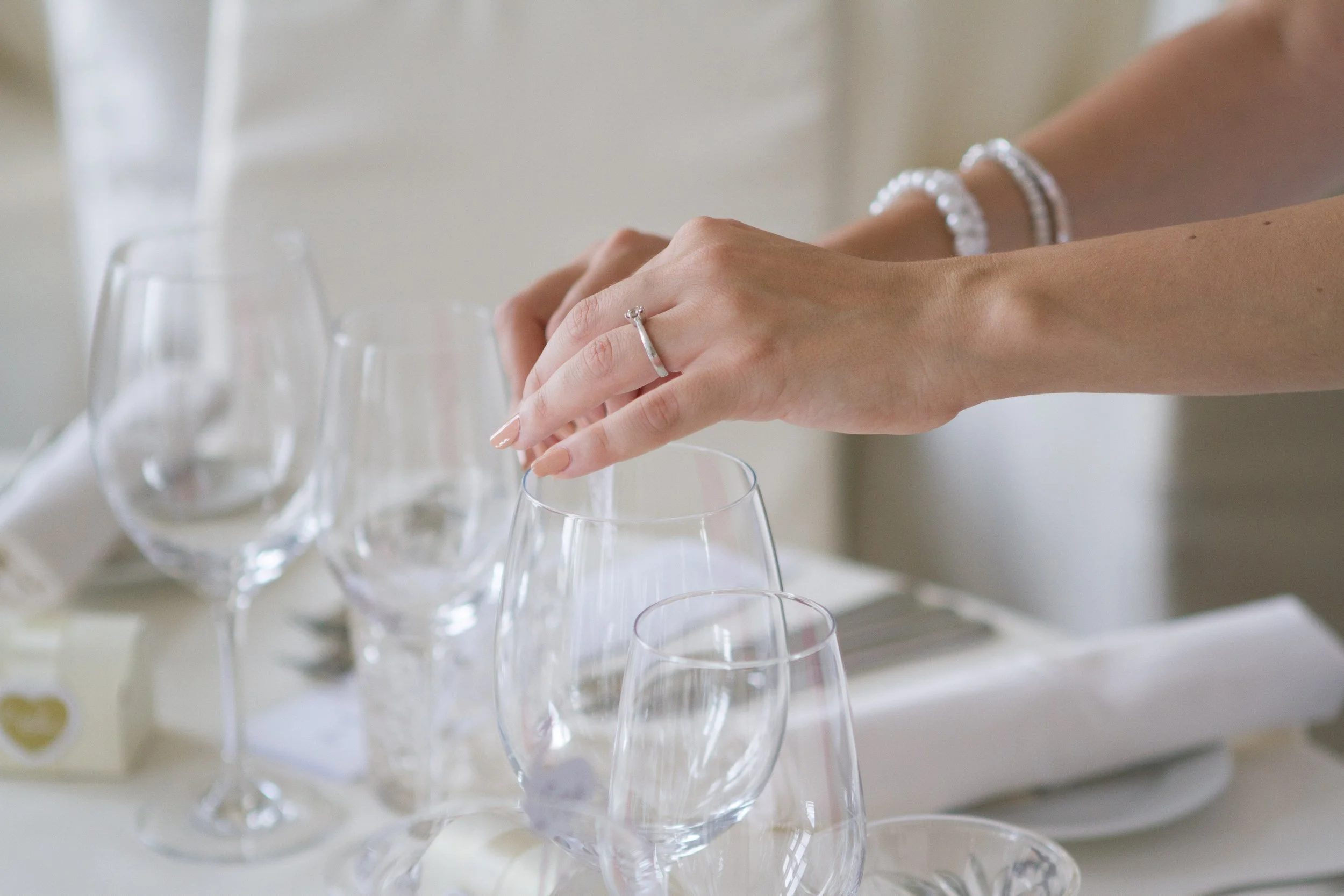 Hand mit Schmuck greift nach einem Weinglas auf einem Tisch, weitere Weingläser sind sichtbar.