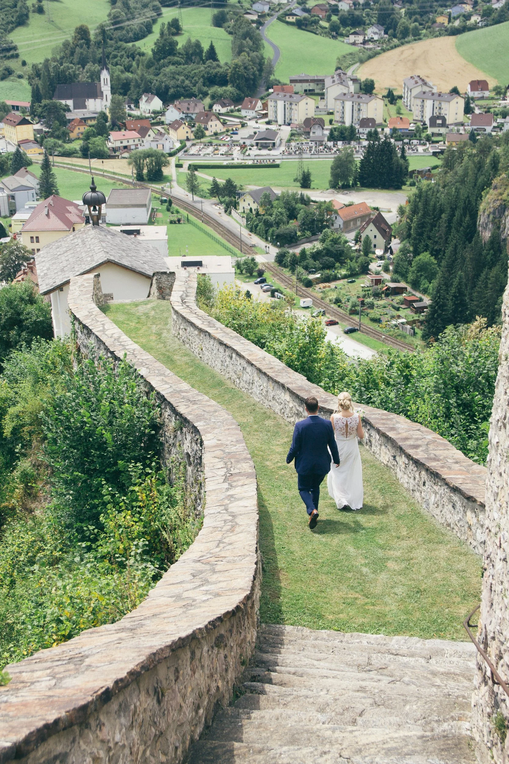 Ein Paar spaziert die Steintreppe hinunter, umgeben von grünen Büschen und Bäumen, mit Blick auf ein kleines Dorf in den Hügeln.