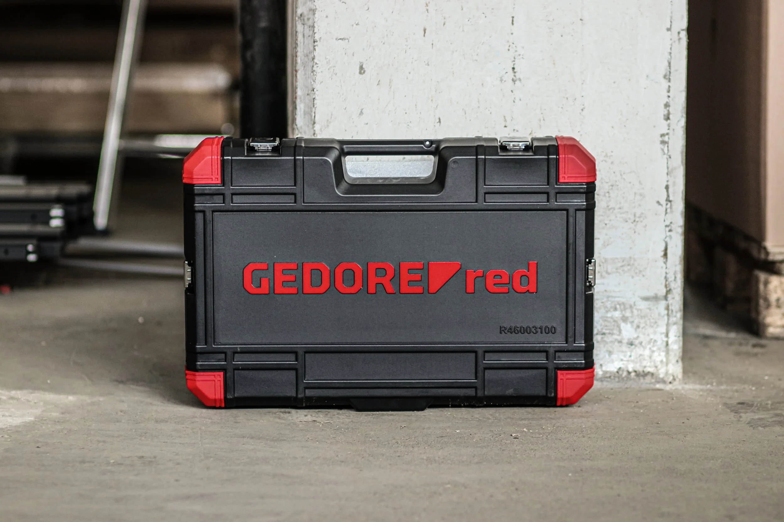 Eine schwarze Werkzeugbox mit roten Ecken und Griff, auf einem Betonboden neben einer weißen Wand.