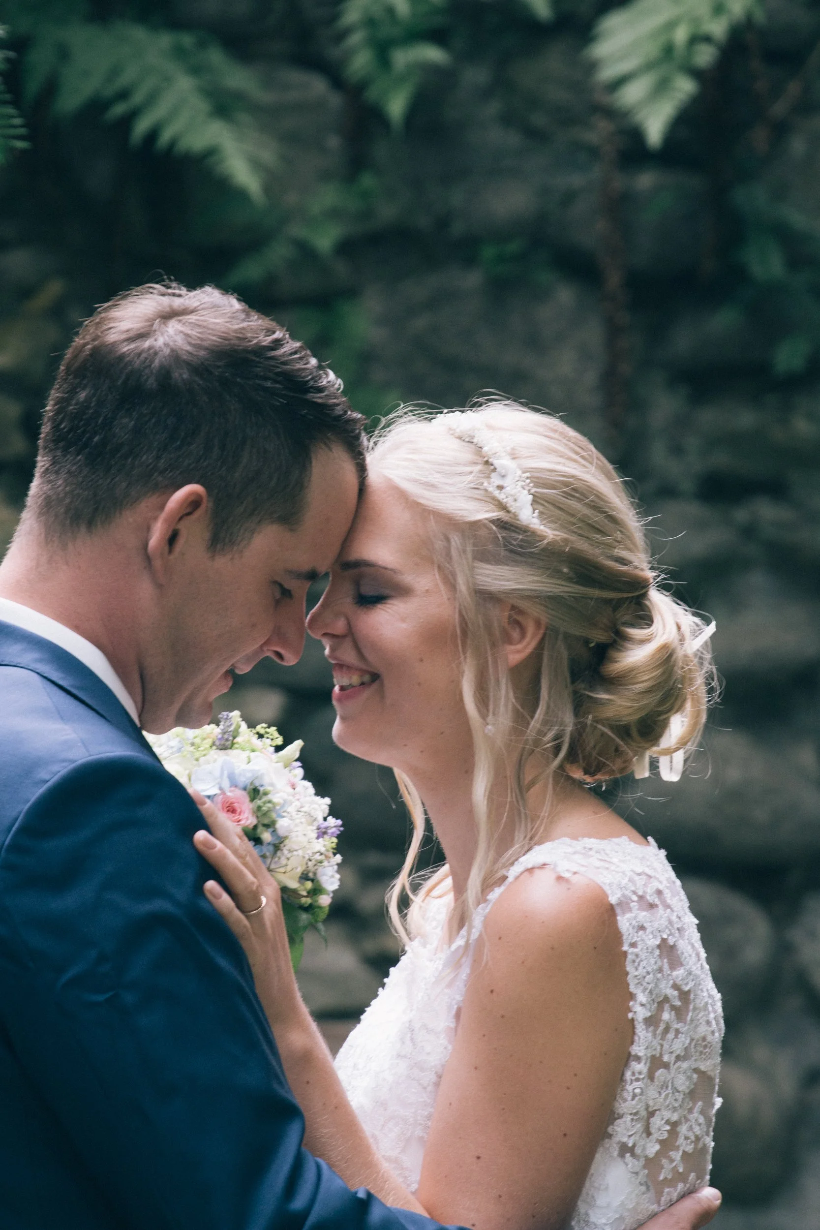 Ein Brautpaar steht sich nahe gegenüber, die Braut trägt ein weißes Spitzenkleid mit einer Schleife im Rücken und eine Blondine mit Hochsteckfrisur, der Bräutigam trägt einen dunklen Anzug, sie sind verliebt und lachen im Garten.