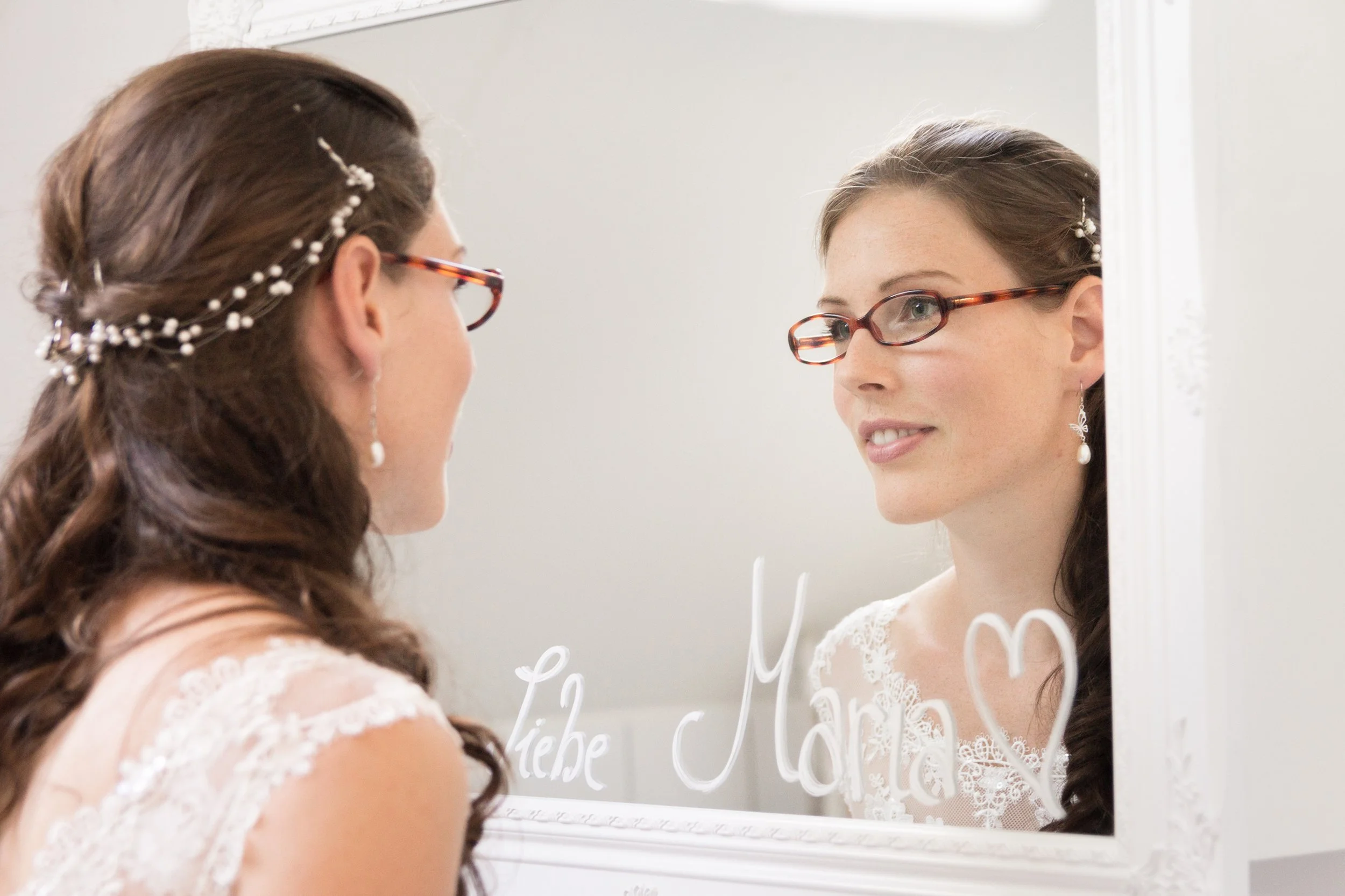 Junge Frau schaut in einen Spiegel, trägt ein Hochzeitskleid, hat lange braune Haare mit Perlen-Accessoires und trägt Brille.