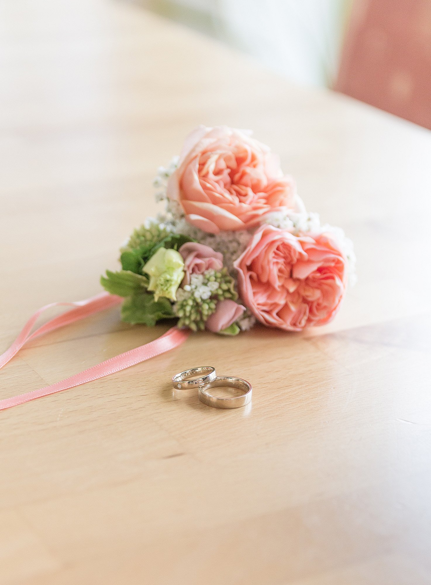 Ein Brautstrauß mit rosa Rosen, kleinen weißen Blumen und grünen Blättern, sowie Eheringe auf einem Holztisch.